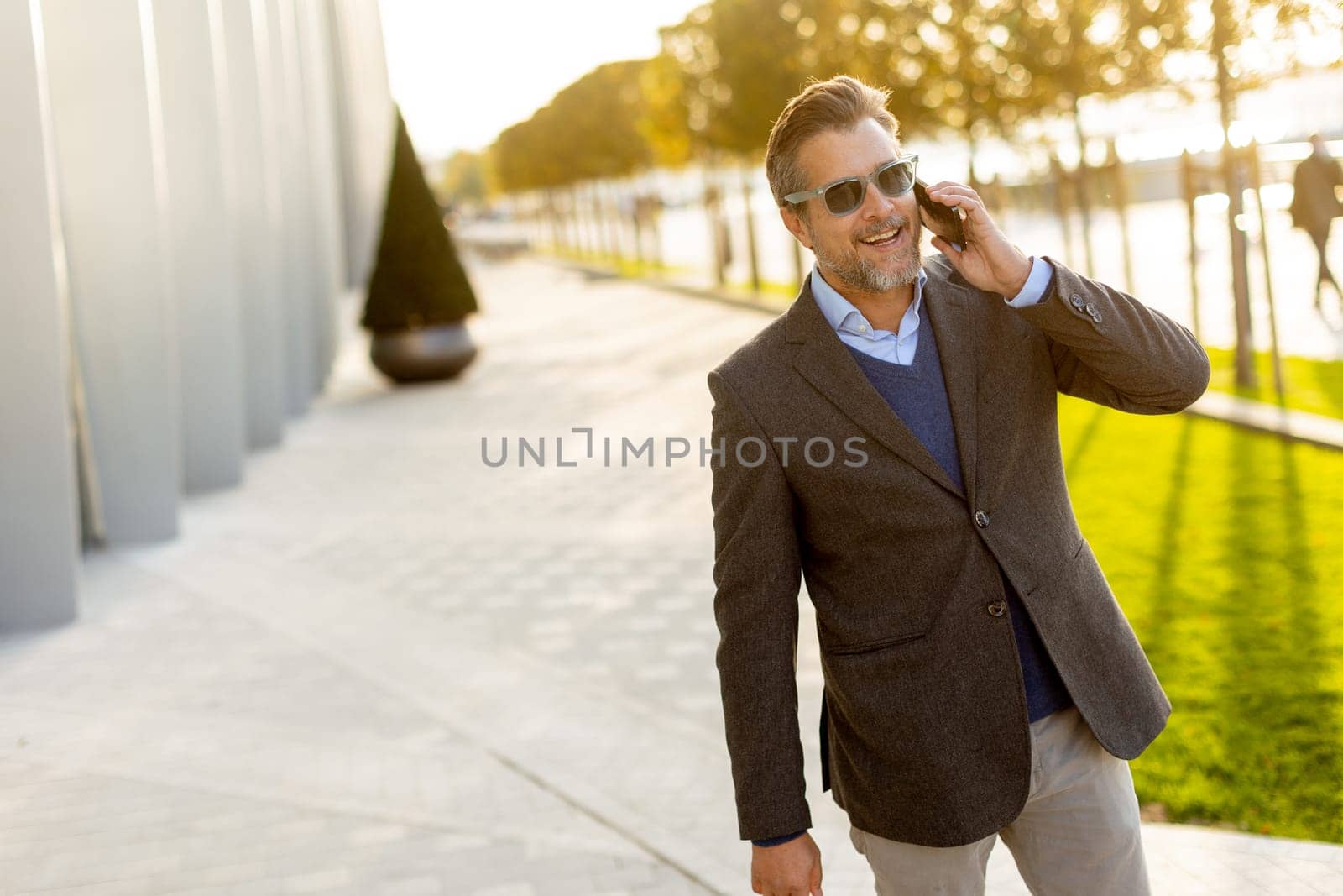 A well-dressed man strolls along a sleek pathway, chatting on his phone under the warm glow of the afternoon sun, surrounded by contemporary architecture and greenery.