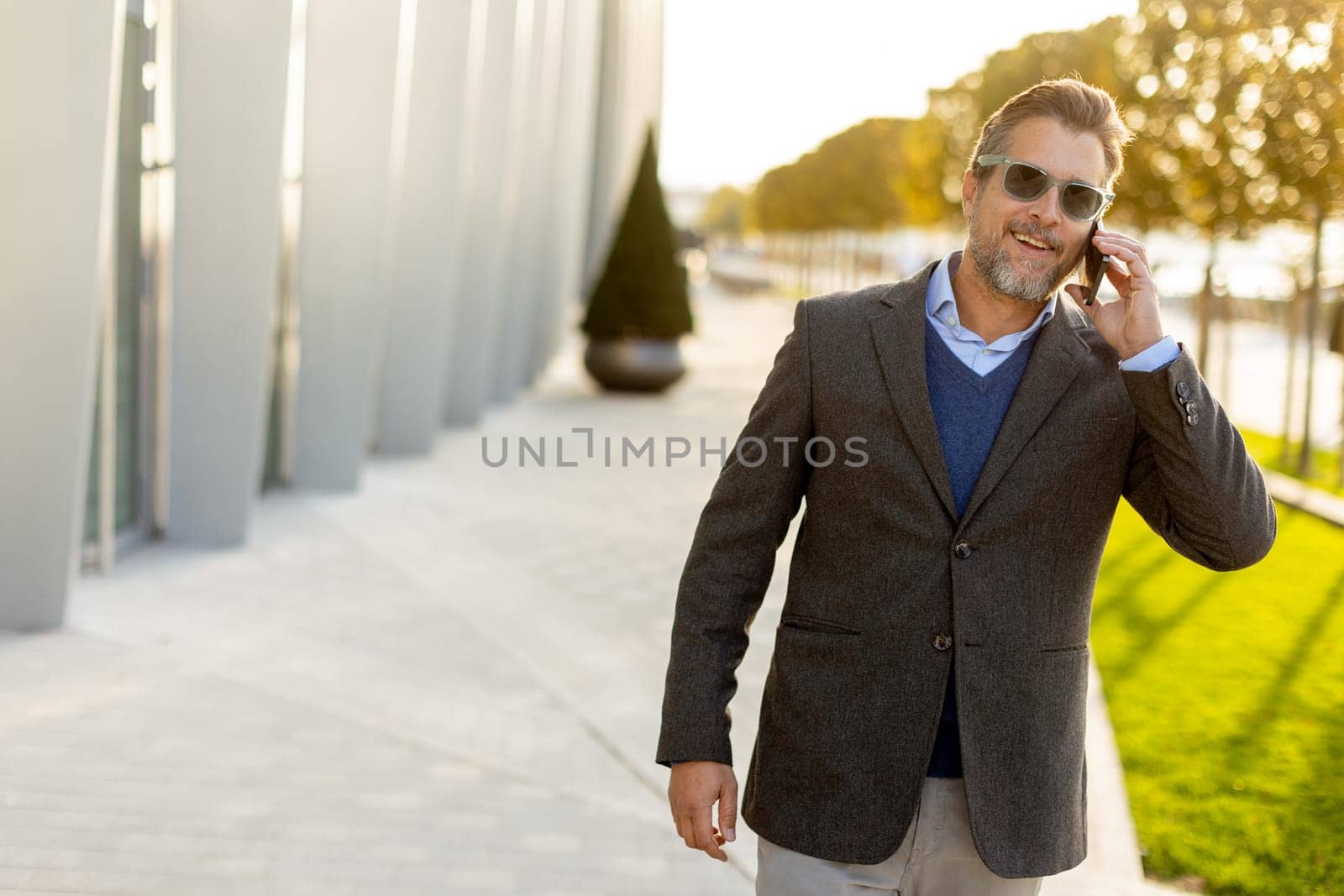 A well-dressed man strolls along a sleek pathway, chatting on his phone under the warm glow of the afternoon sun, surrounded by contemporary architecture and greenery.