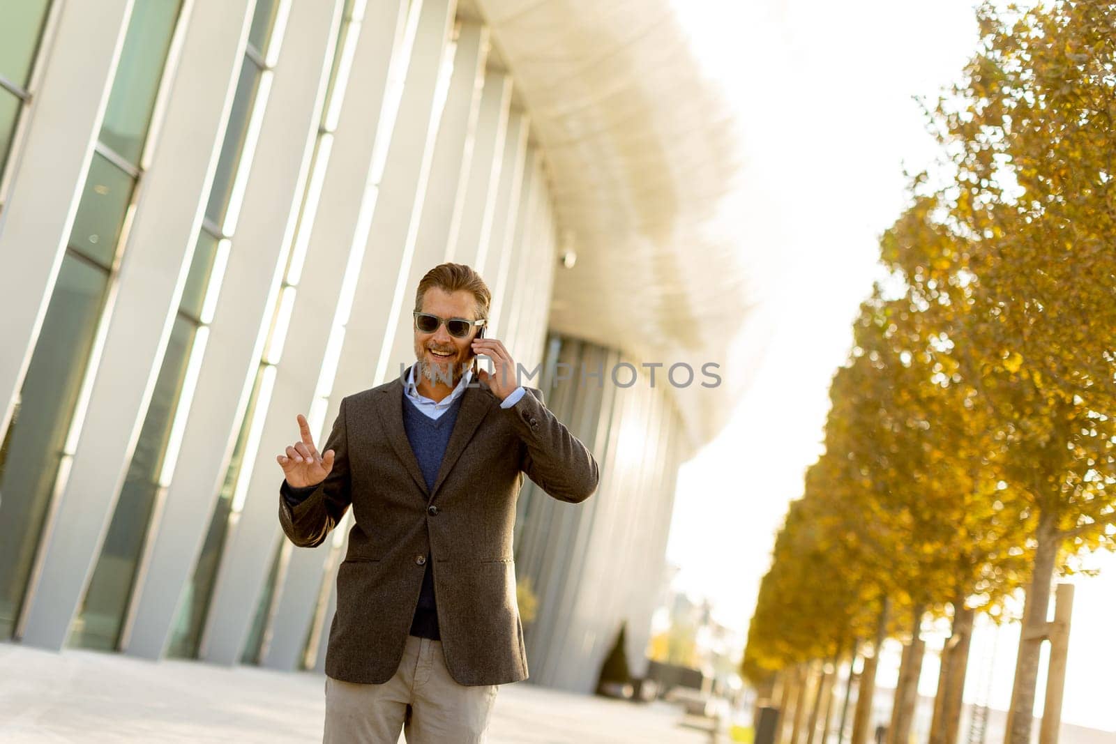 A well-dressed man strolls along a sleek pathway, chatting on his phone under the warm glow of the afternoon sun, surrounded by contemporary architecture and greenery.