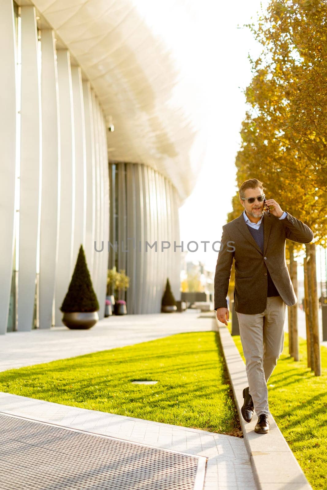 A well-dressed man strolls along a sleek pathway, chatting on his phone under the warm glow of the afternoon sun, surrounded by contemporary architecture and greenery.
