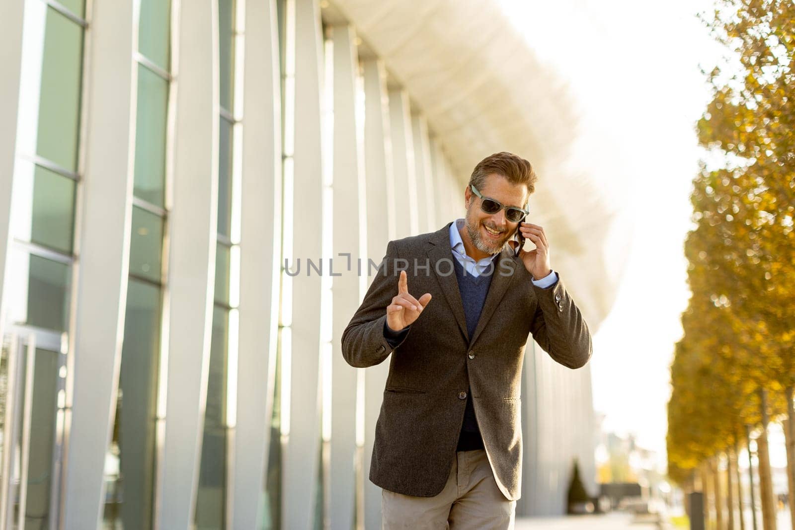 A well-dressed man strolls along a sleek pathway, chatting on his phone under the warm glow of the afternoon sun, surrounded by contemporary architecture and greenery.