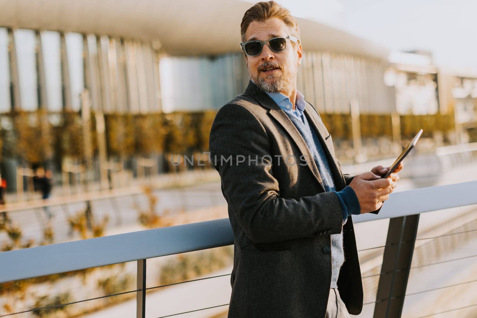 An elegant individual in a blazer stands on a balcony at sunset, holding a tablet and gazing over a vibrant metropolis, embodying professionalism and relaxation.