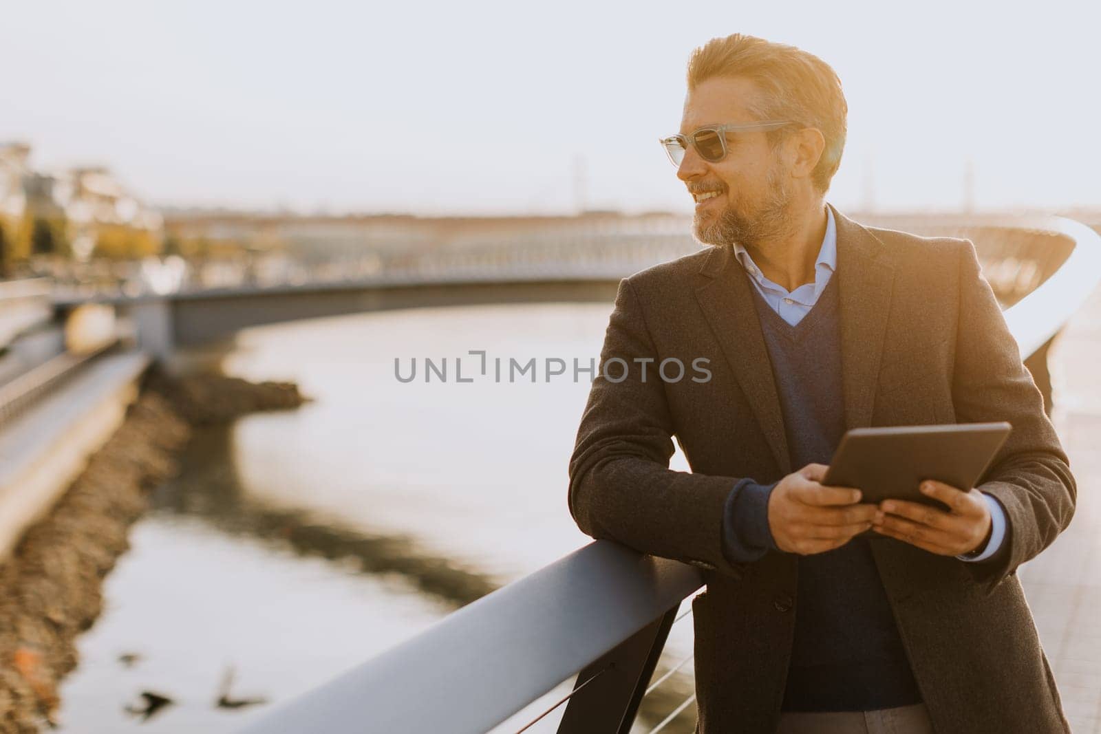Wearing a stylish outfit, a man with a warm smile leans against a railing by the river at sunset, engaging with his tablet in a peaceful and picturesque environment.