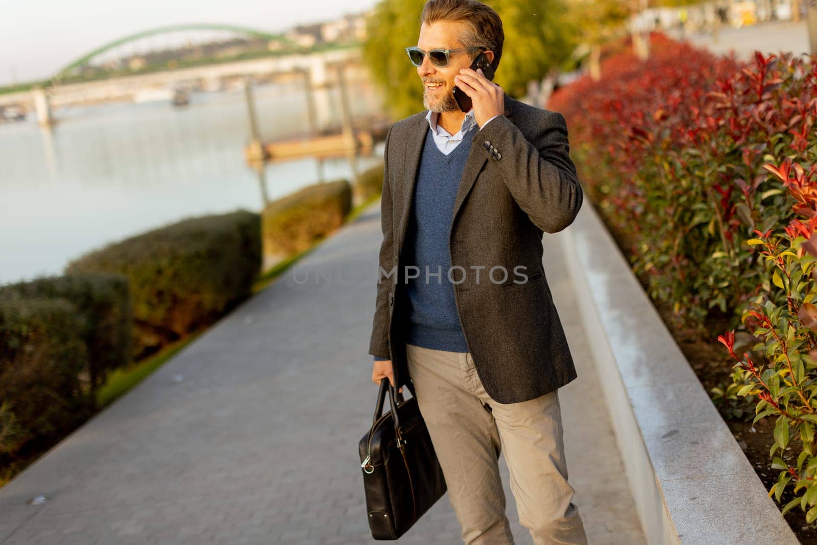 A well-dressed man strolls along a riverside walkway while talking on his phone. The vibrant plants and calming water create a serene atmosphere perfect for business discussions.