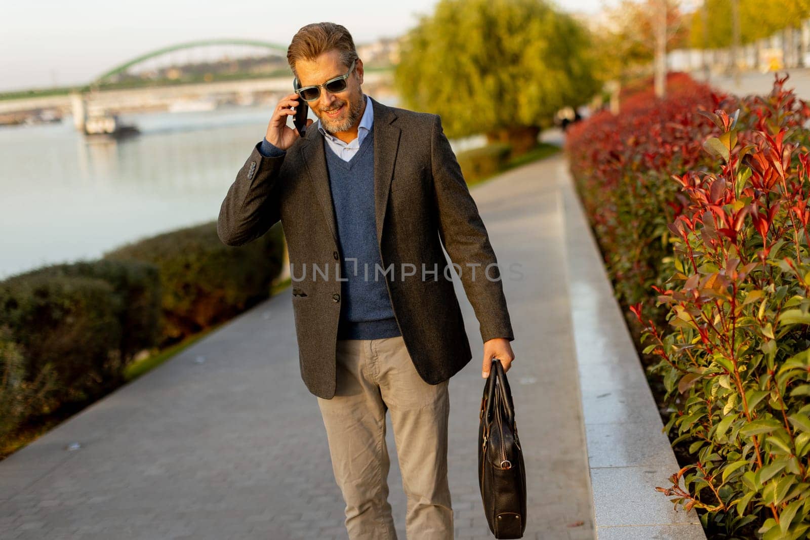 Dressed in a smart casual outfit, a man walks confidently by the water, engaged in a phone conversation while admiring the vibrant autumn colors around him