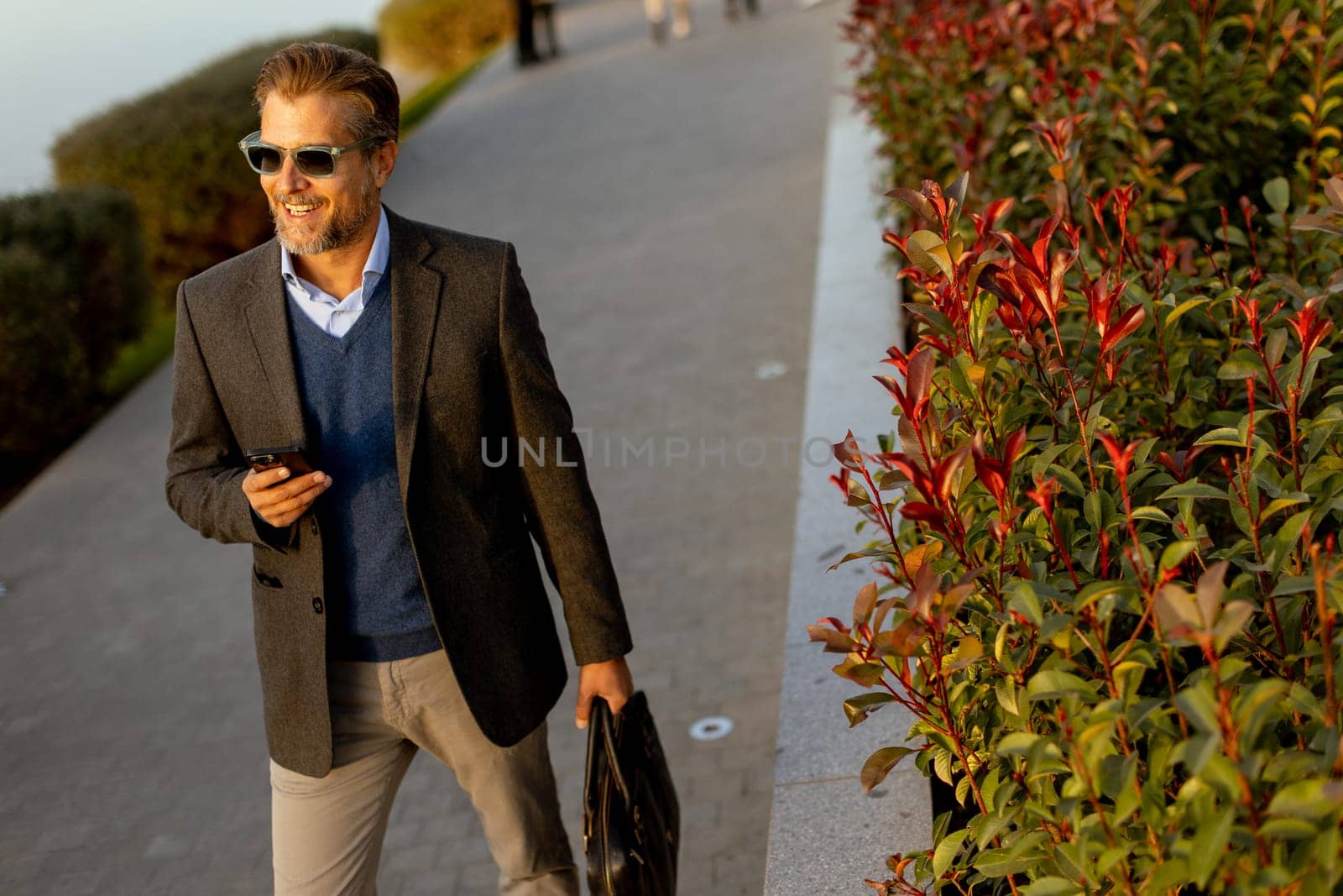 A well-dressed man walks confidently along a picturesque waterfront path, checking his phone, with colorful leaves and a serene backdrop accentuating the tranquil golden hour.