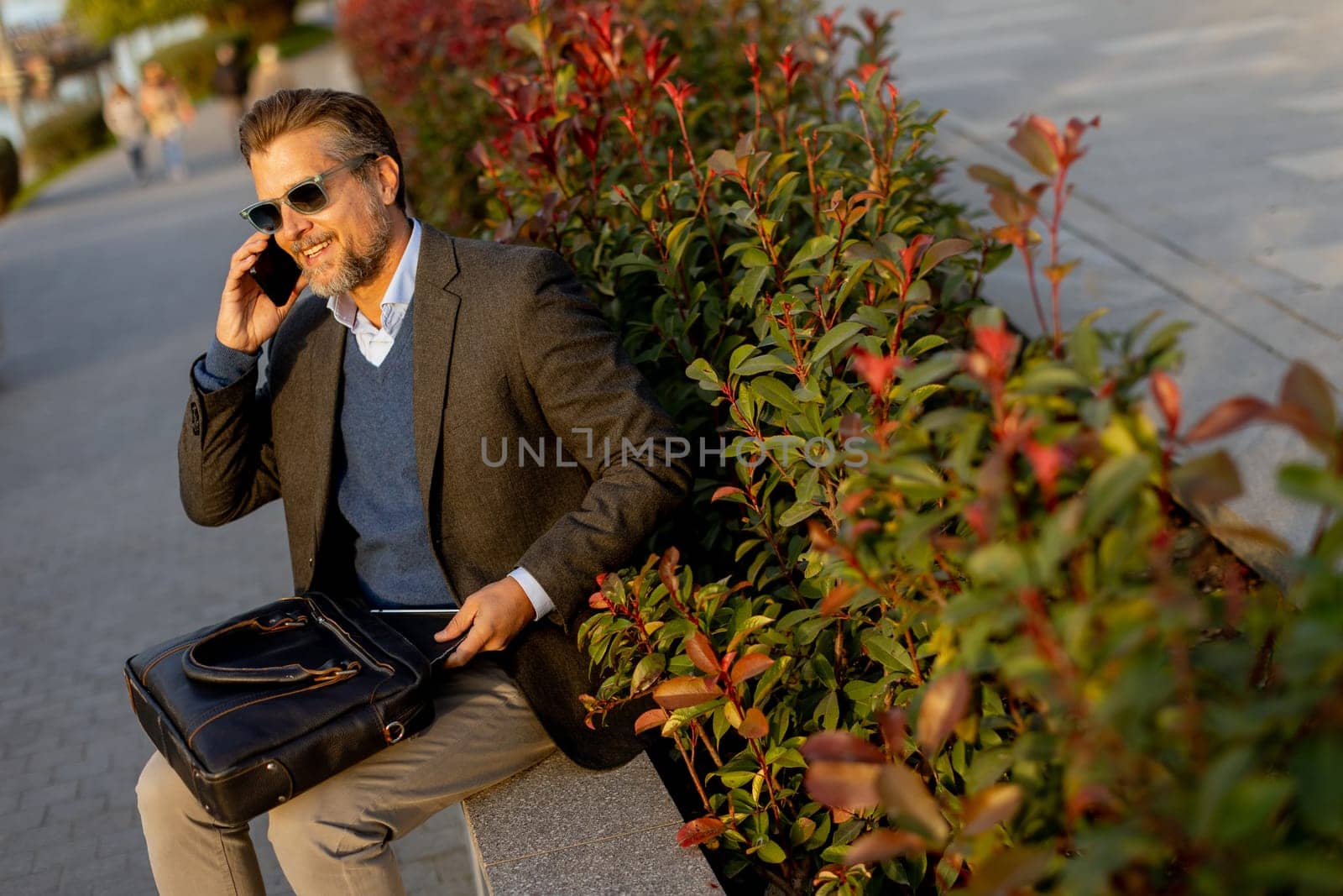 A professional man dressed in a blazer enjoys a sunny afternoon, chatting on his phone beside lush plants while carrying a leather bag. The scene reflects a blend of work and relaxation.