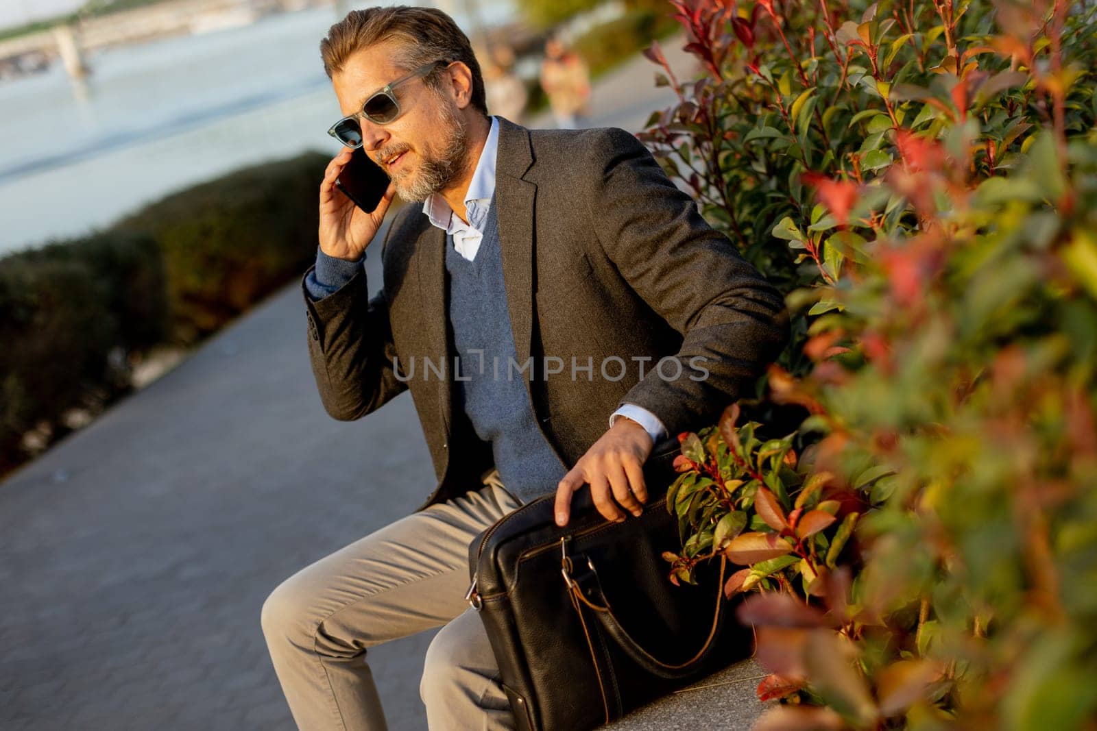 A professional man dressed in a blazer enjoys a sunny afternoon, chatting on his phone beside lush plants while carrying a leather bag. The scene reflects a blend of work and relaxation.