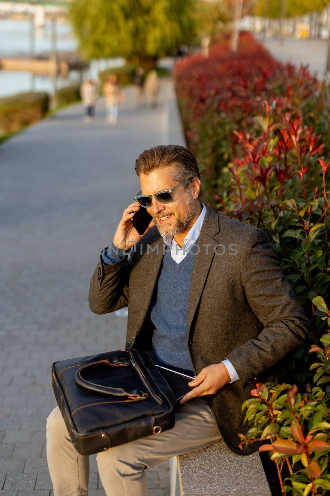A professional man dressed in a blazer enjoys a sunny afternoon, chatting on his phone beside lush plants while carrying a leather bag. The scene reflects a blend of work and relaxation.
