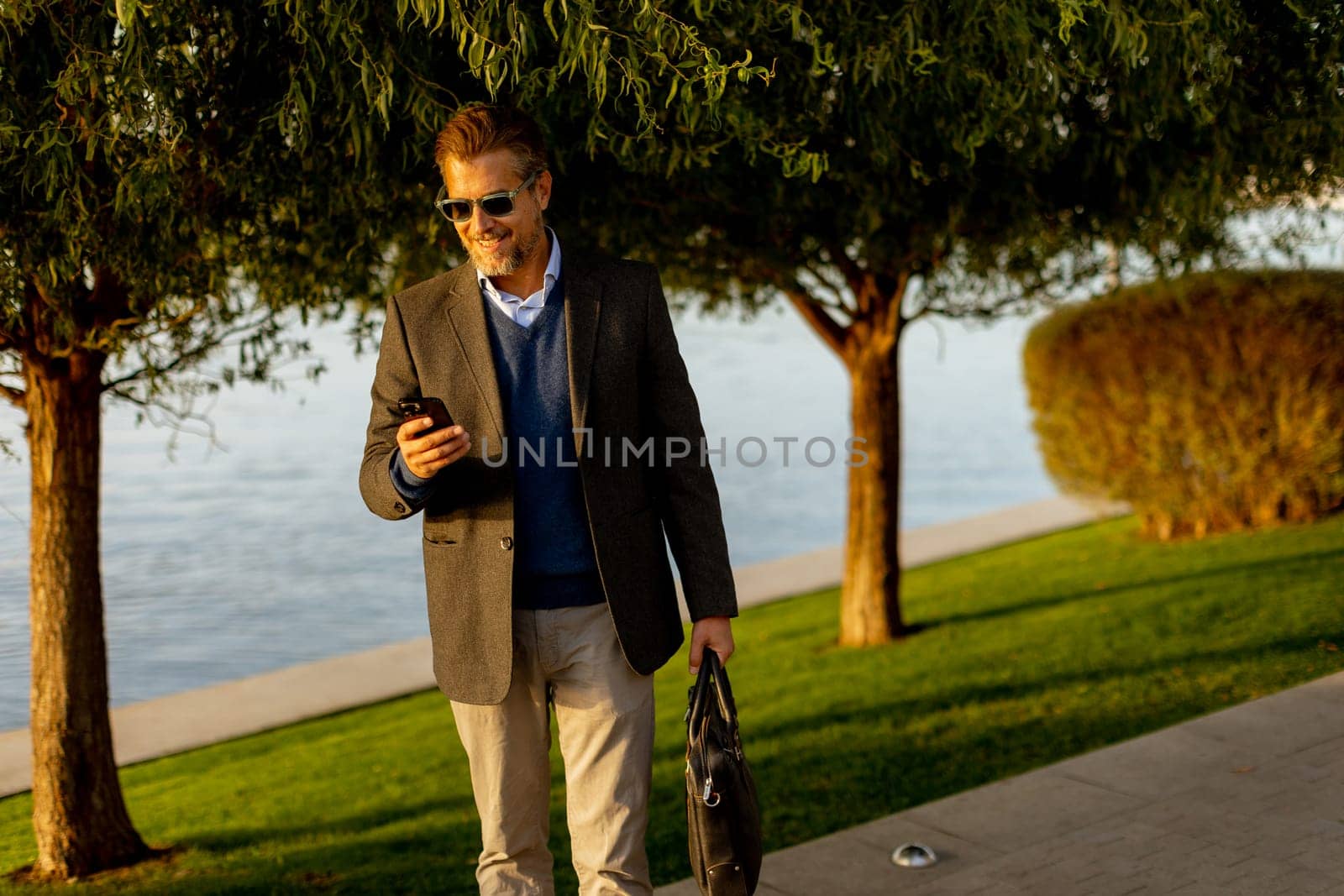 Dressed in a smart casual outfit, a man walks confidently by the water, engaged in a phone conversation while admiring the vibrant autumn colors around him