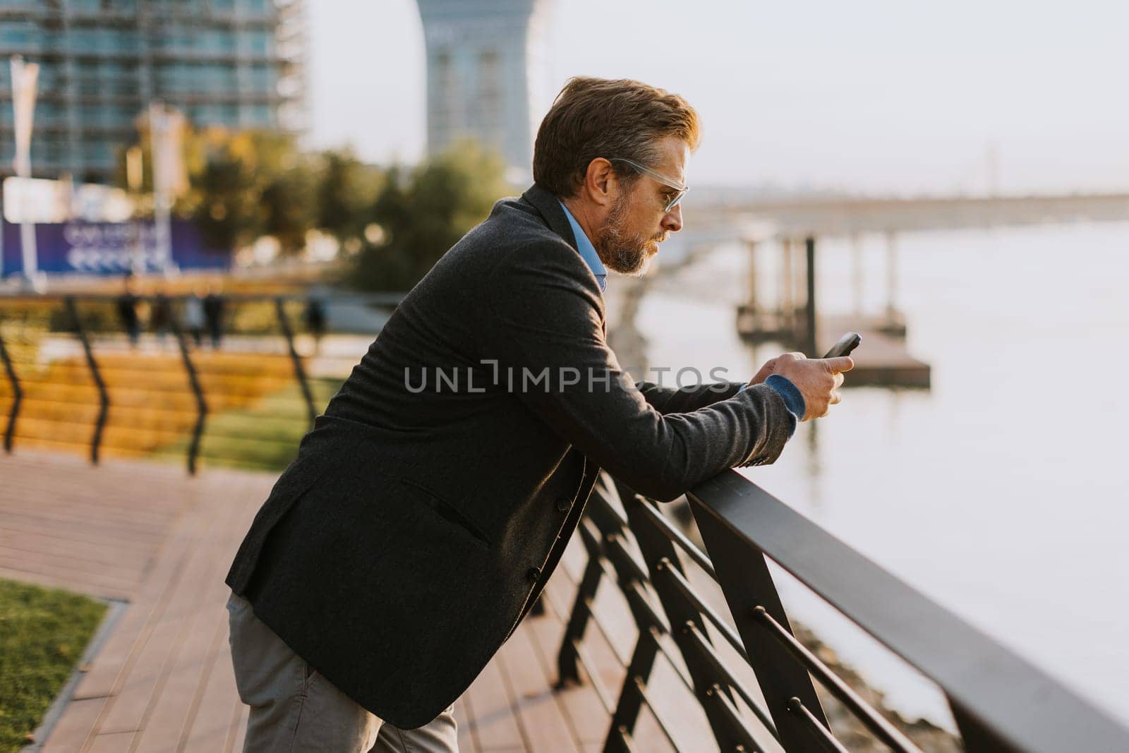 A relaxed man leans on a railing, enjoying a phone conversation as the sun sets over the water, casting a warm glow over the serene riverside surroundings.