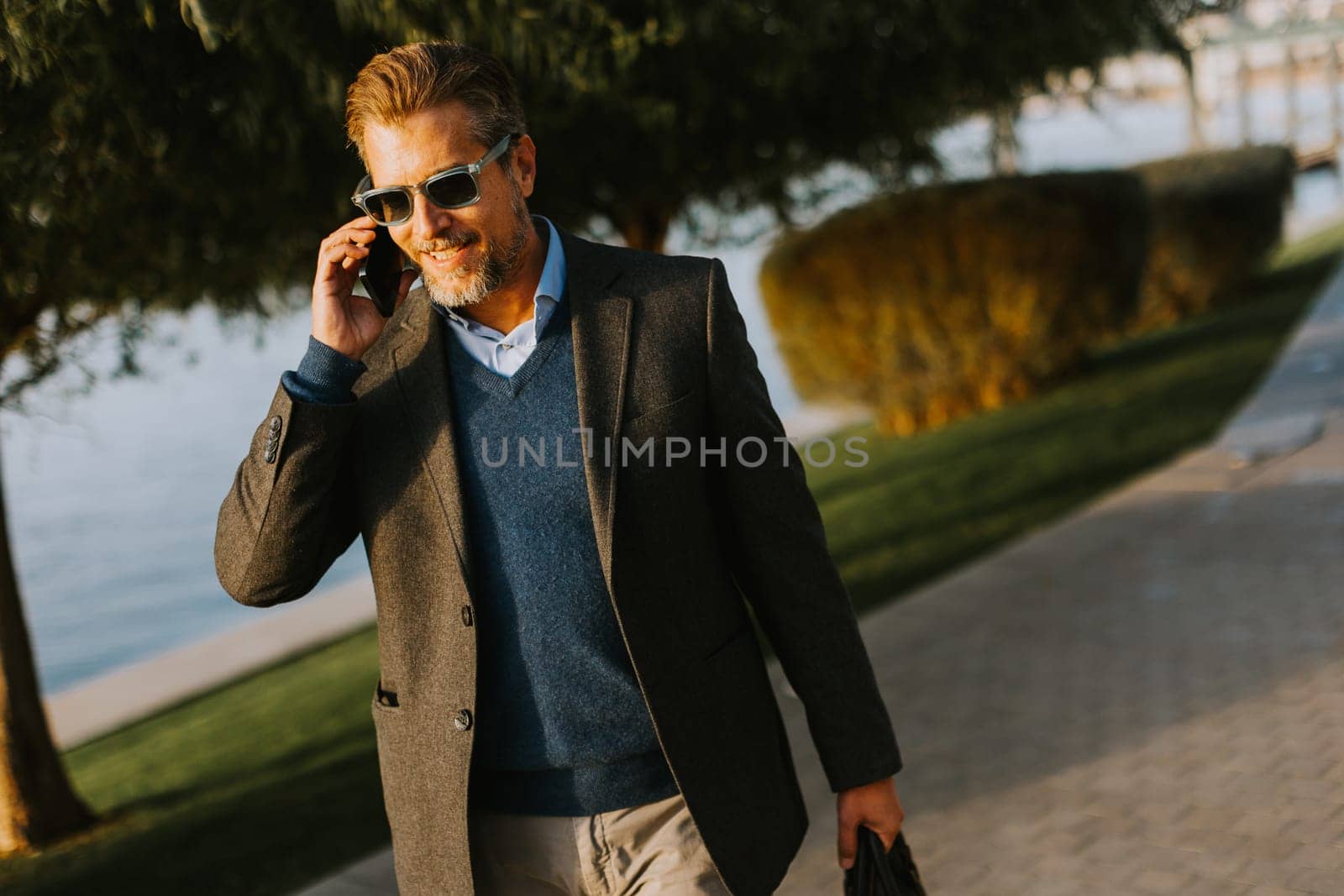 A well-dressed man strolls by the water, absorbed in a phone call. Surrounded by lush greenery, he pairs sophistication with casual ease on a sunny day.