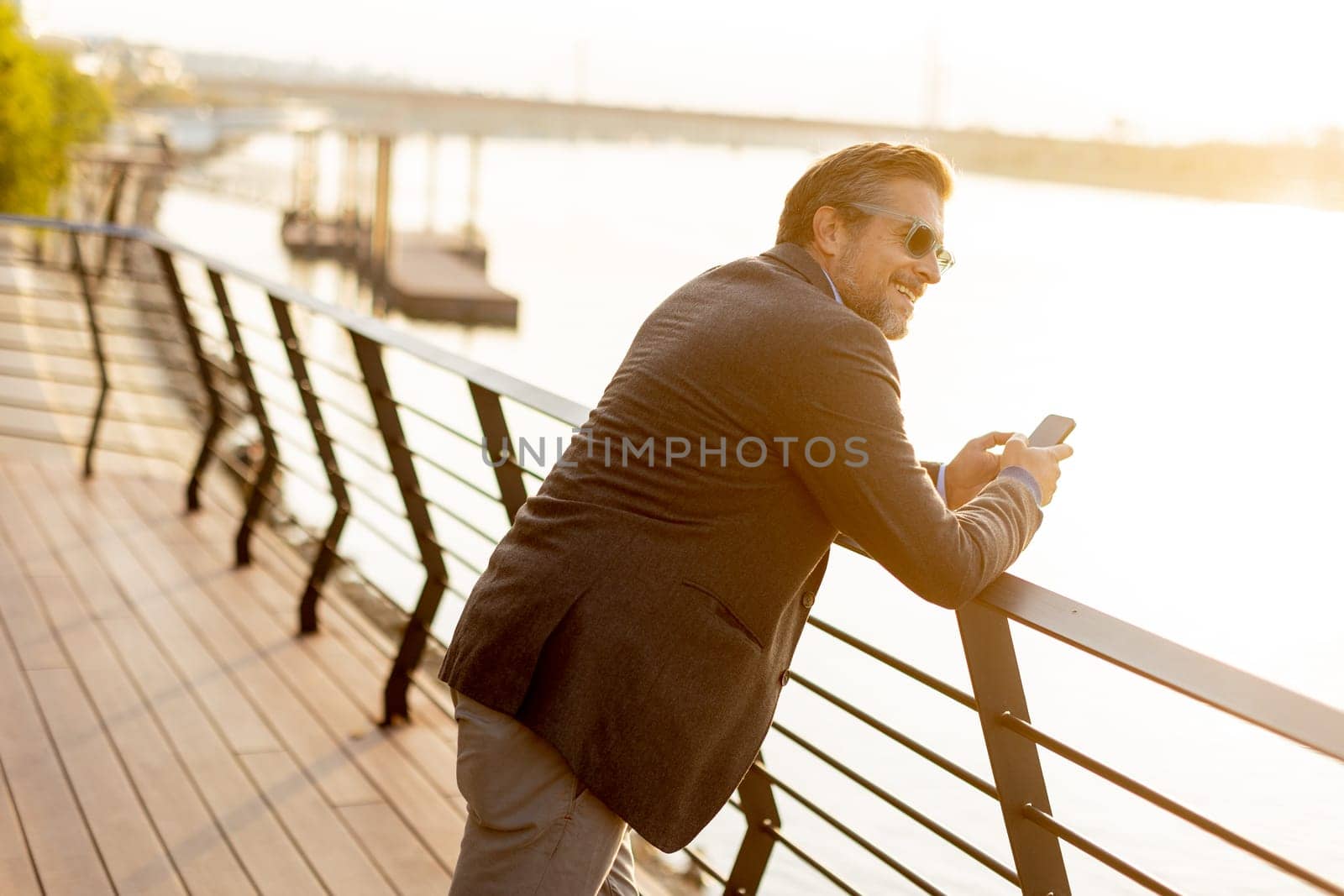 A relaxed man leans on a railing, enjoying a phone conversation as the sun sets over the water, casting a warm glow over the serene riverside surroundings.