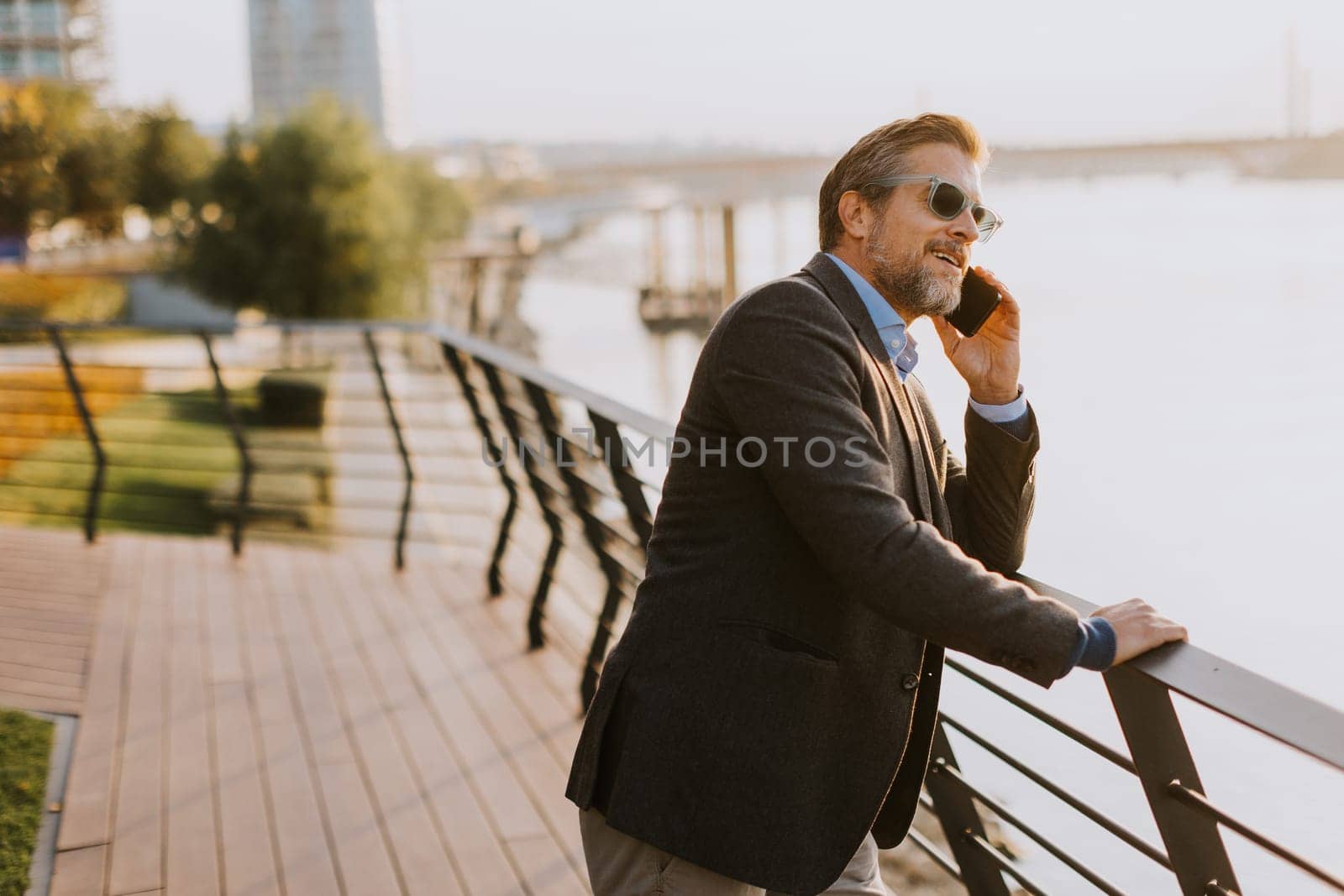 A relaxed man leans on a railing, enjoying a phone conversation as the sun sets over the water, casting a warm glow over the serene riverside surroundings.