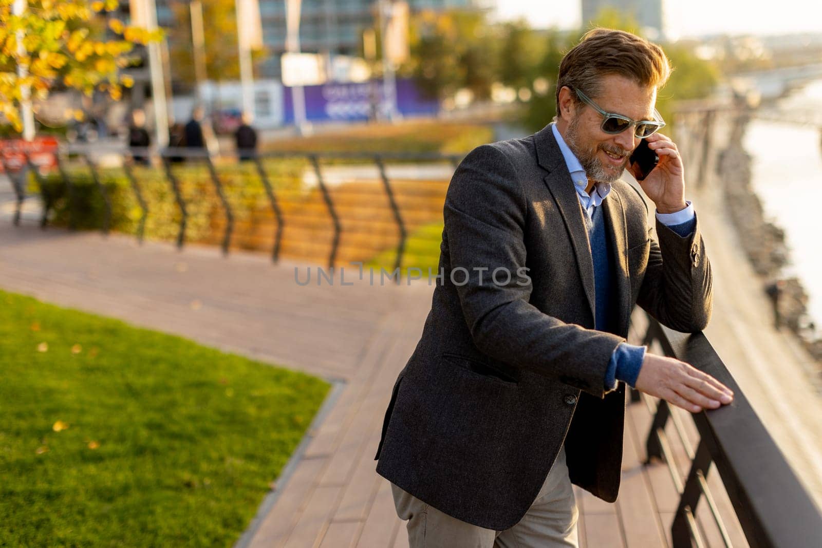 Dressed in smart casual attire, a man stands by an urban landscape, engaging in a phone conversation as autumn leaves swirl around him in the warm sunlight.