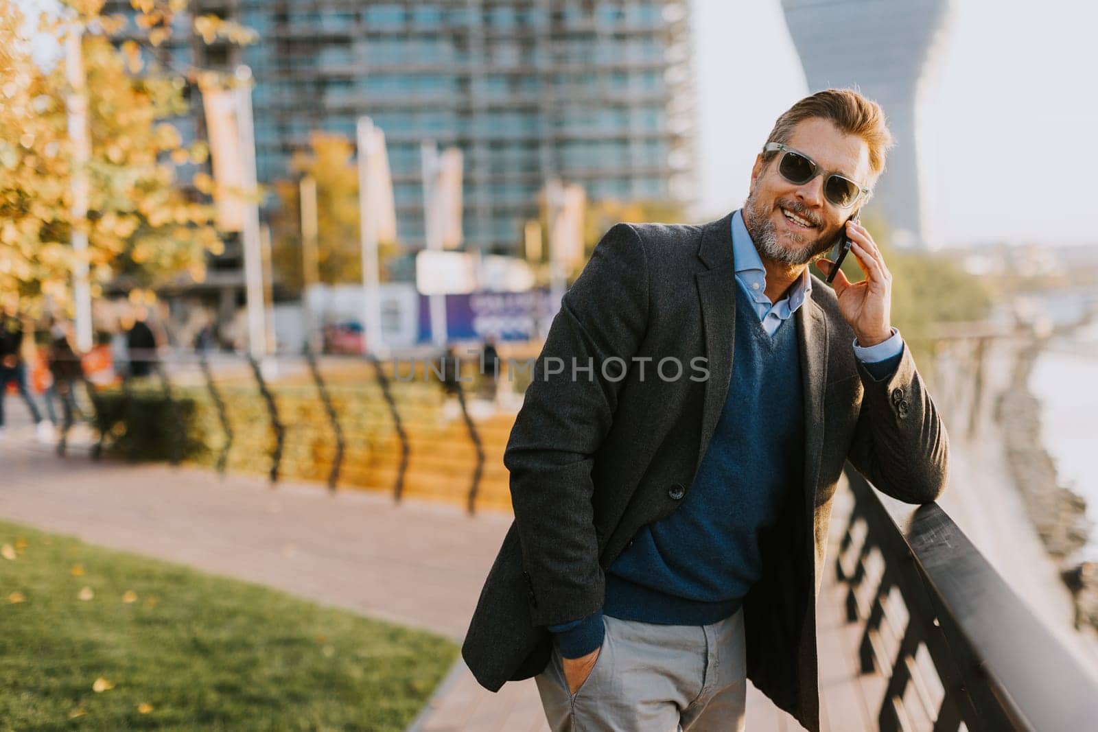 Dressed in smart casual attire, a man stands by an urban landscape, engaging in a phone conversation as autumn leaves swirl around him in the warm sunlight.