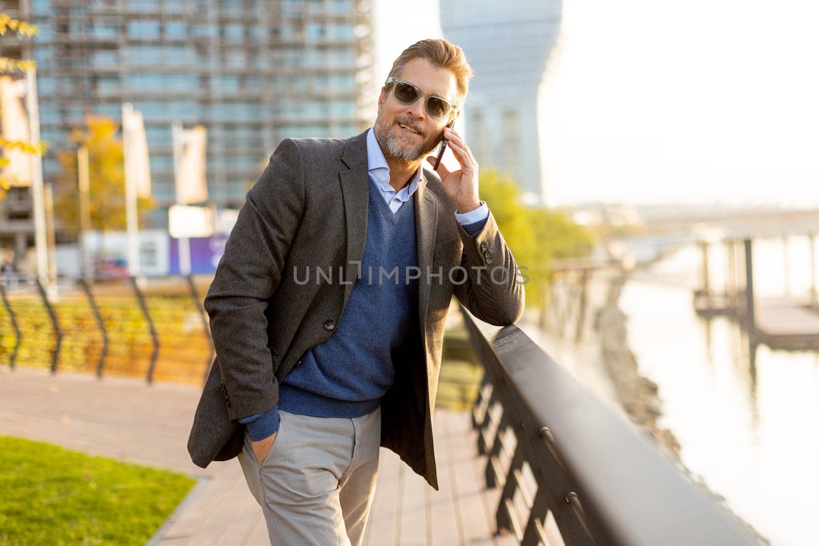 Dressed in smart casual attire, a man stands by an urban landscape, engaging in a phone conversation as autumn leaves swirl around him in the warm sunlight.