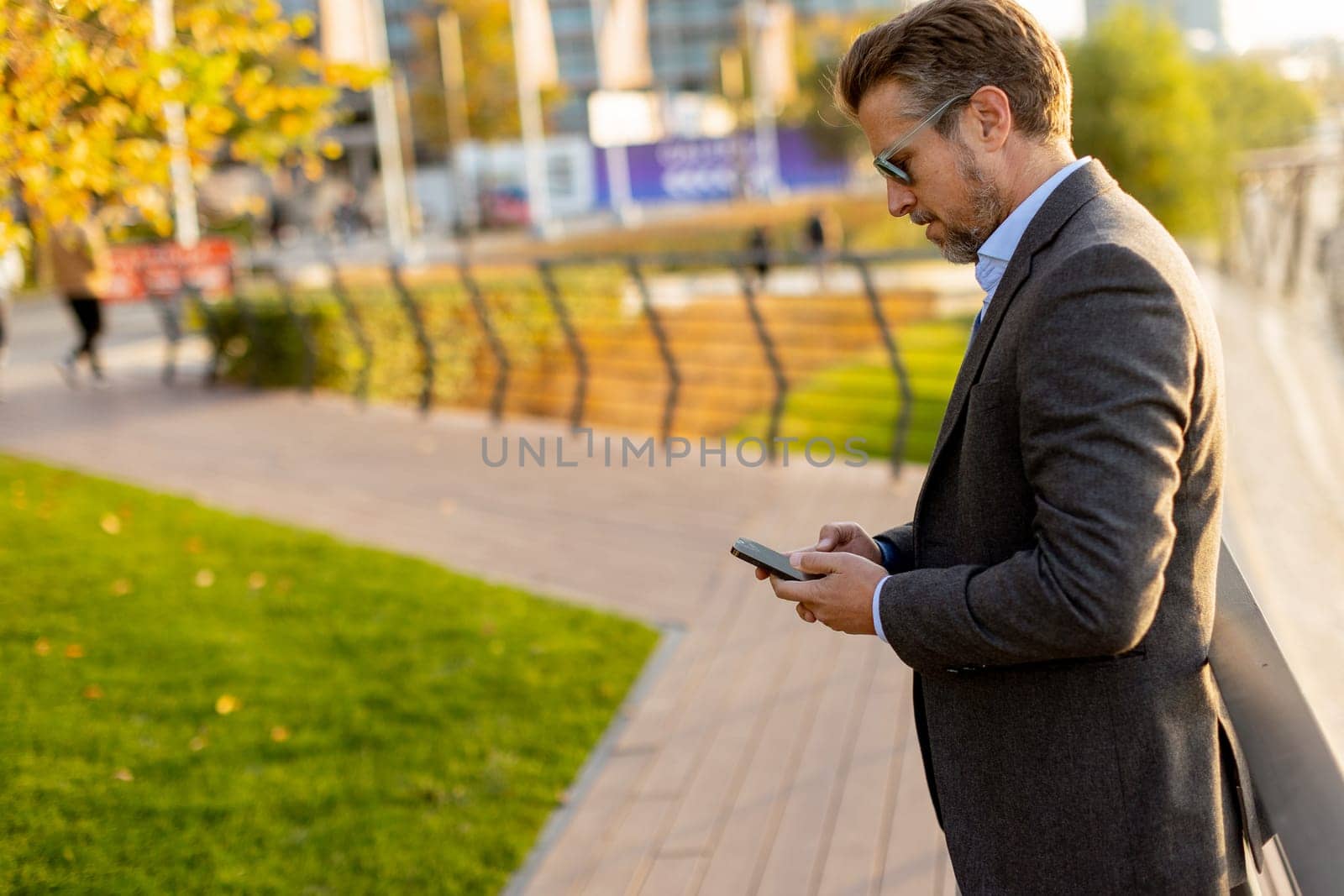Dressed in smart casual attire, a man stands by an urban landscape, engaging in a phone conversation as autumn leaves swirl around him in the warm sunlight.