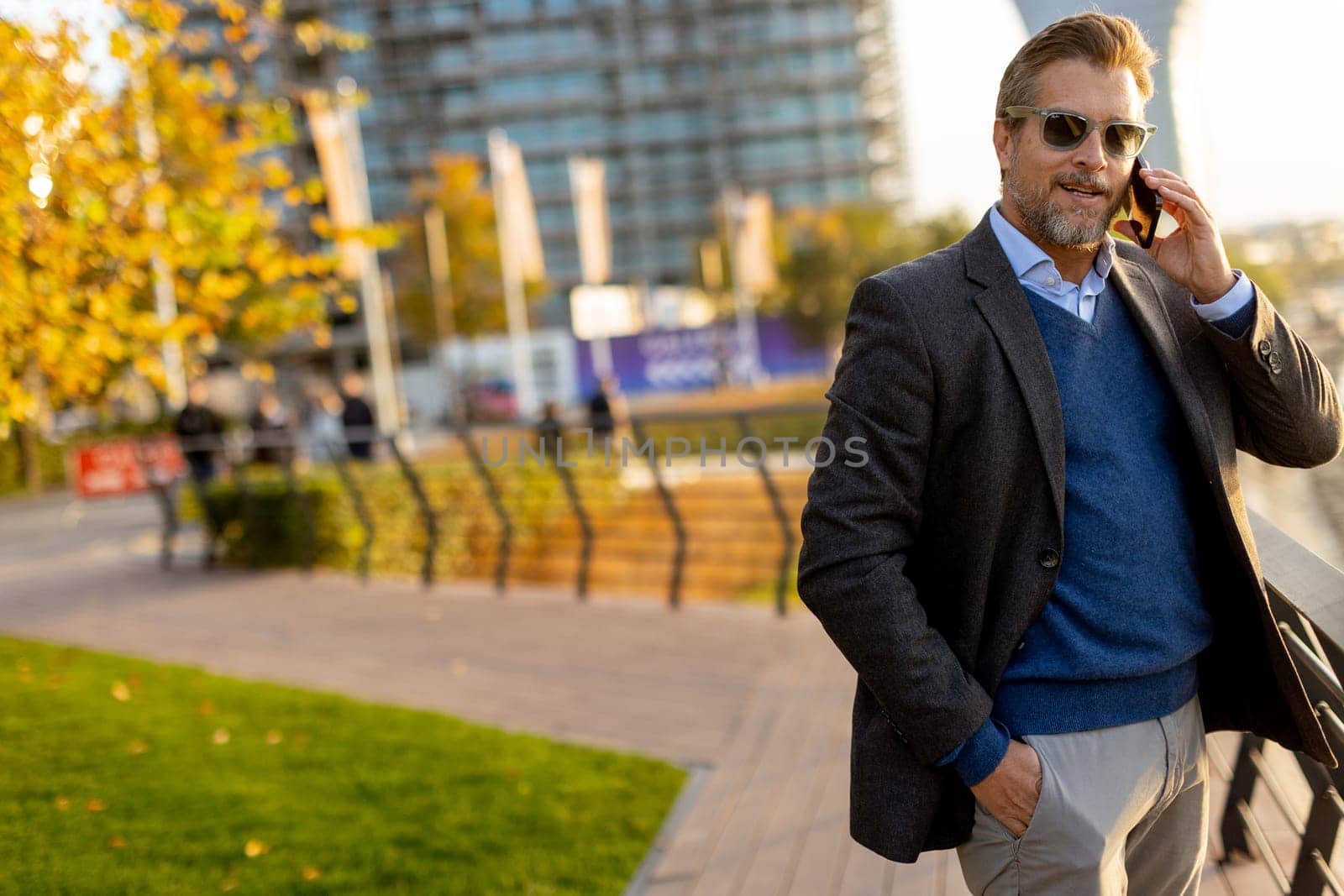 Dressed in smart casual attire, a man stands by an urban landscape, engaging in a phone conversation as autumn leaves swirl around him in the warm sunlight.