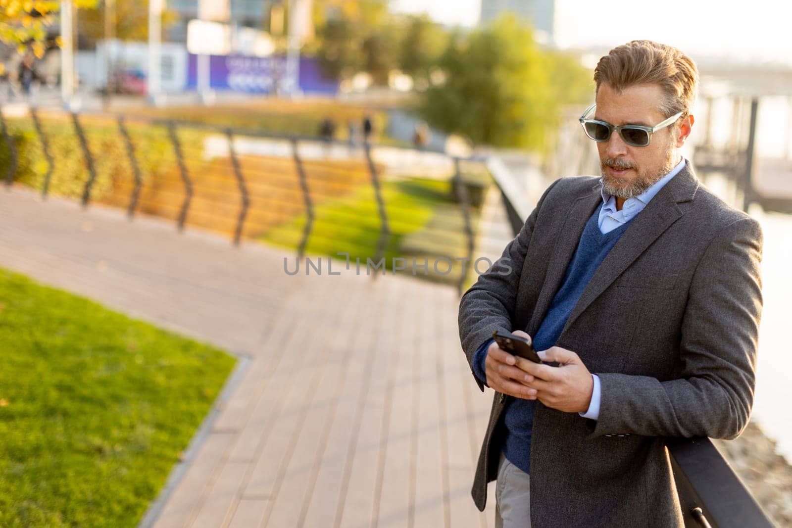 Dressed in smart casual attire, a man stands by an urban landscape, engaging in a phone conversation as autumn leaves swirl around him in the warm sunlight.
