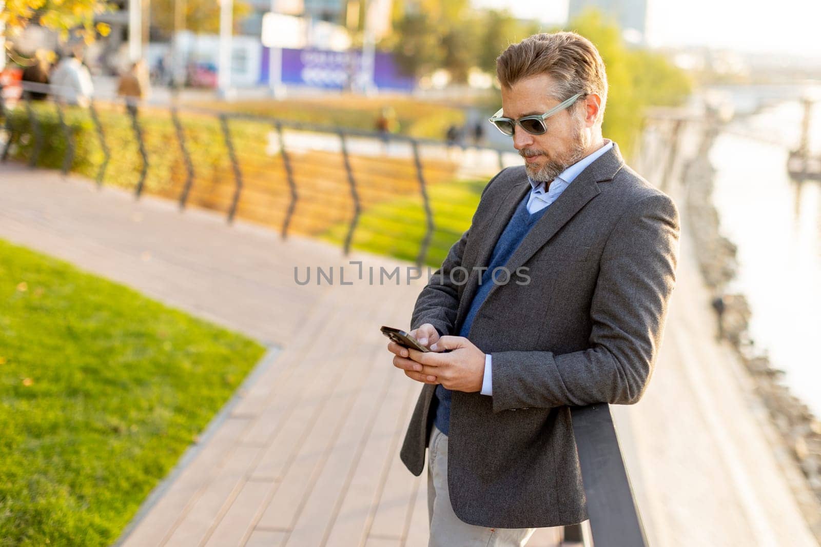 Dressed in smart casual attire, a man stands by an urban landscape, engaging in a phone conversation as autumn leaves swirl around him in the warm sunlight.