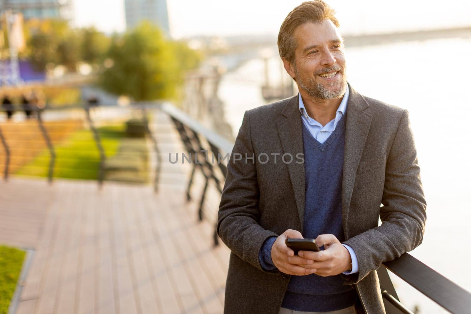 A well-dressed man takes a leisurely walk along the riverside park, engaging with his smartphone as golden hour light casts a warm glow over the landscape.