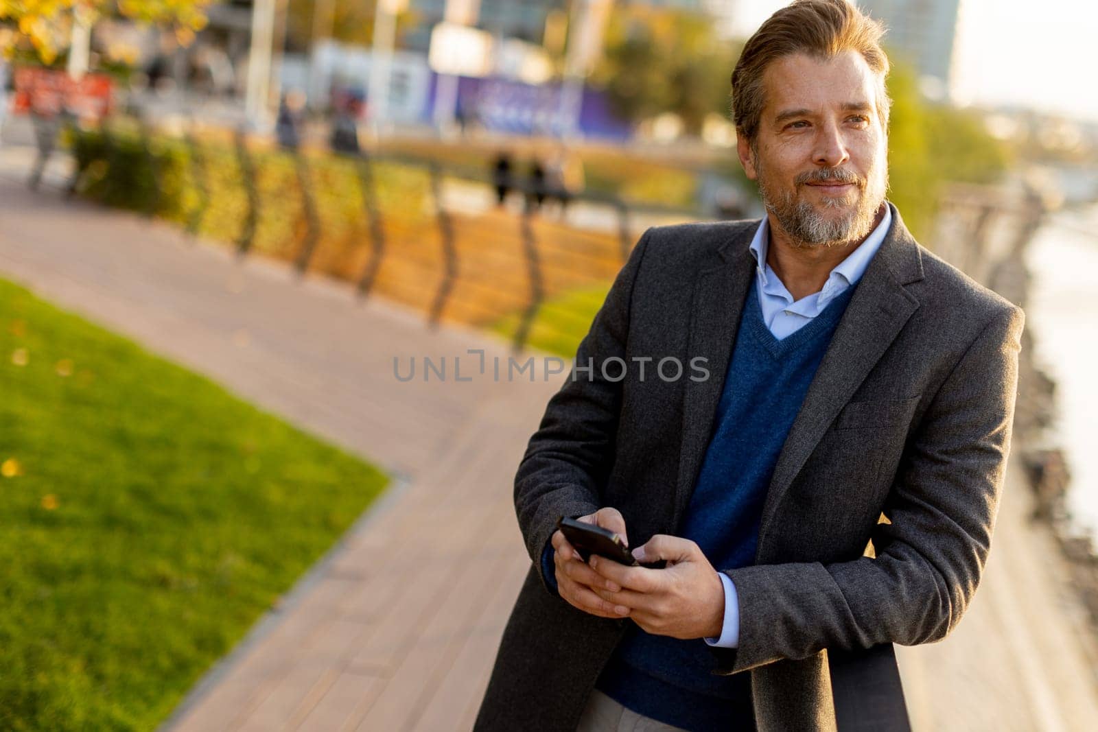 A well-dressed man takes a leisurely walk along the riverside park, engaging with his smartphone as golden hour light casts a warm glow over the landscape.