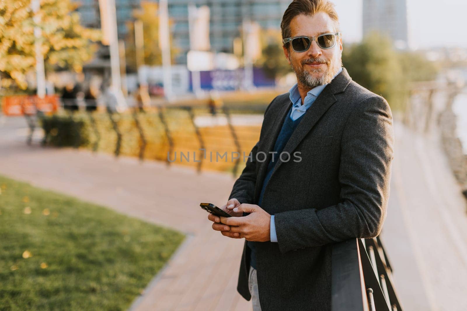 Dressed in smart casual attire, a man stands by an urban landscape, engaging in a phone conversation as autumn leaves swirl around him in the warm sunlight.