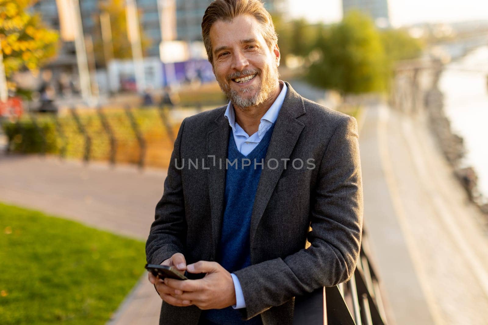 A well-dressed man takes a leisurely walk along the riverside park, engaging with his smartphone as golden hour light casts a warm glow over the landscape.