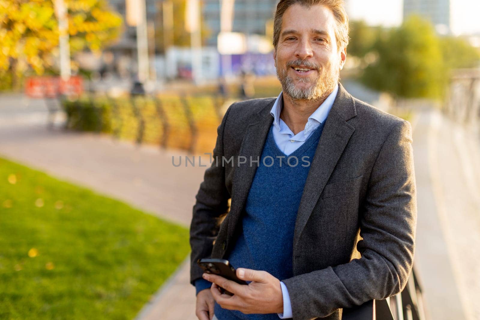 Under the warm sunlight, a relaxed professional is casually dressed, engaging with his smartphone amidst a lively outdoor environment filled with greenery and city life.