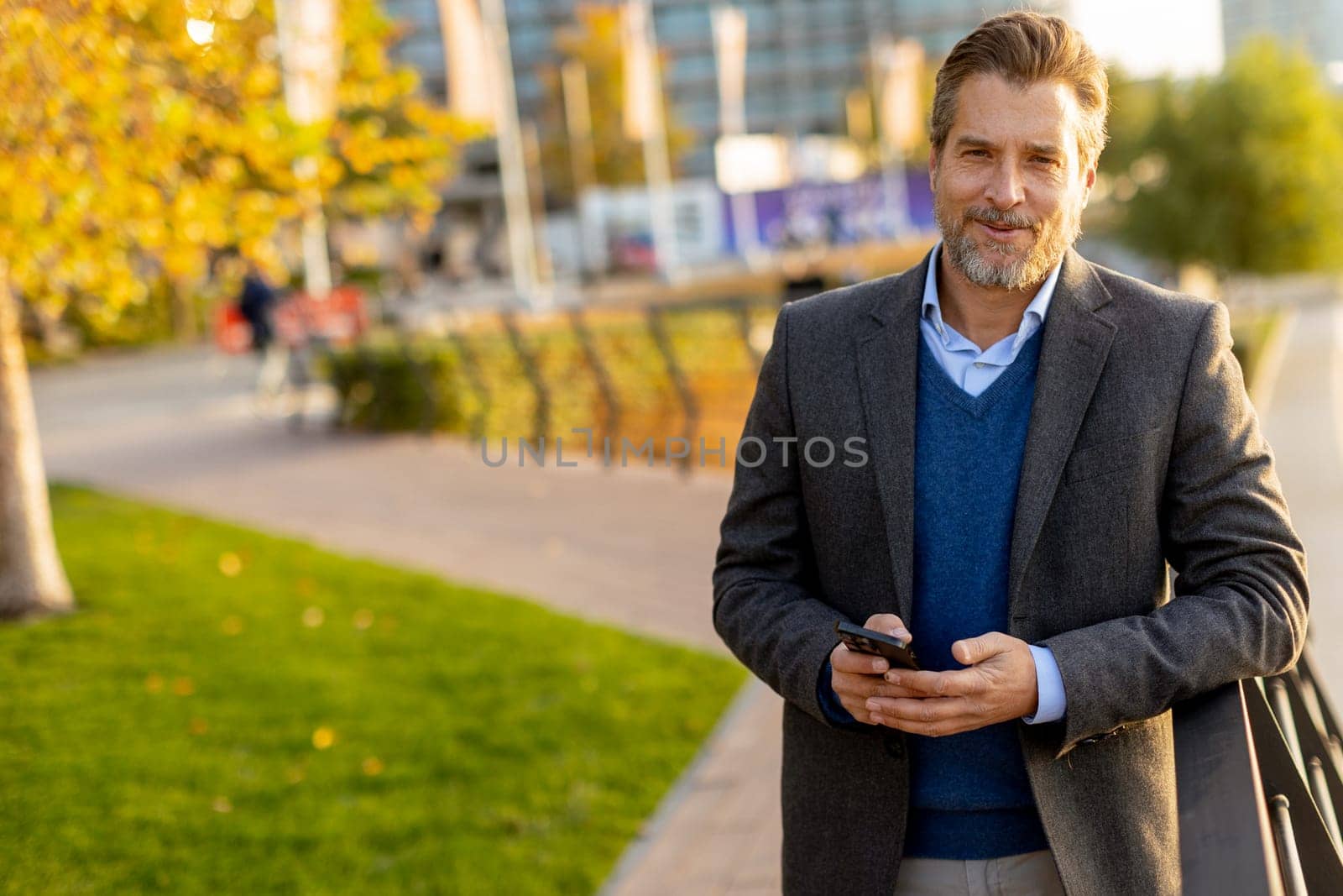 Under the warm sunlight, a relaxed professional is casually dressed, engaging with his smartphone amidst a lively outdoor environment filled with greenery and city life.