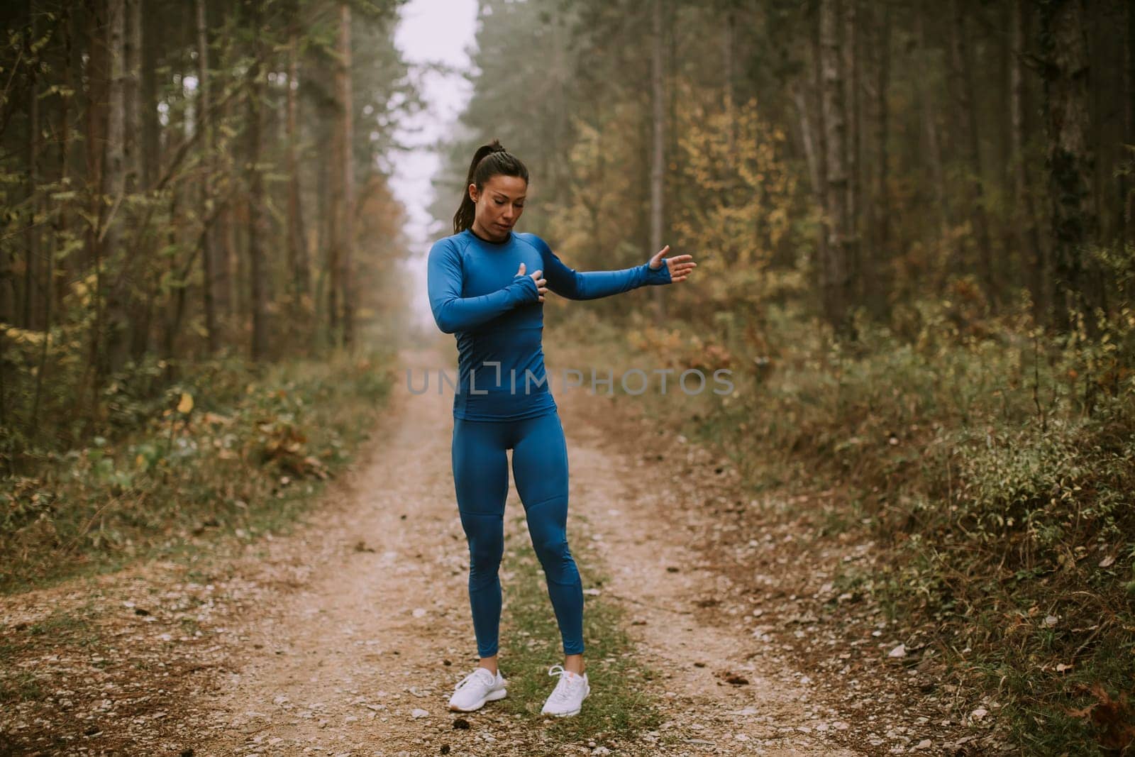 Pretty young woman in blue track suit stretching before workout in the autumn forest