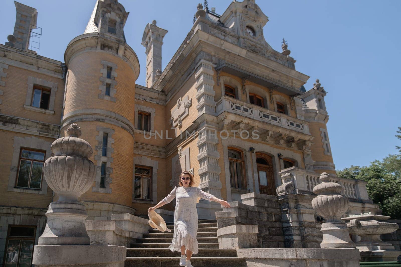 A woman is standing on a set of stairs in front of a large building. She is wearing a white dress and a straw hat. The building has a lot of architectural details, including a clock on the side