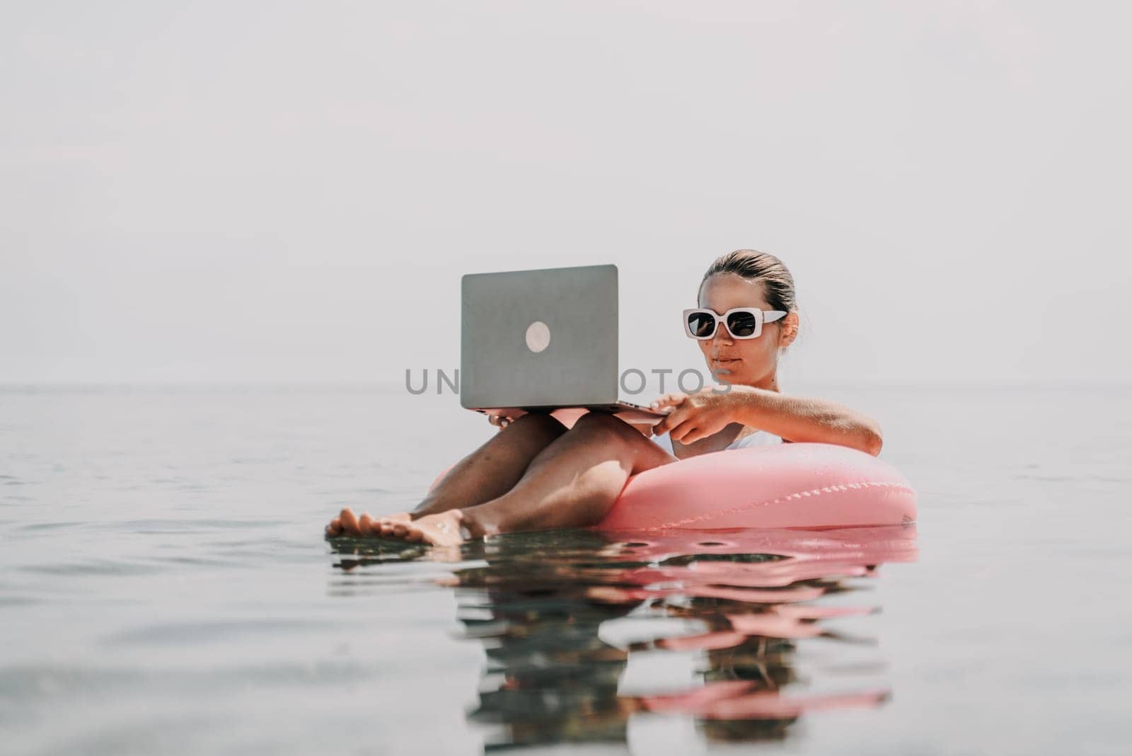 A woman is sitting on a pink inflatable raft in the ocean, using a laptop. Concept of relaxation and leisure, as the woman is enjoying her time in the water while working on her laptop
