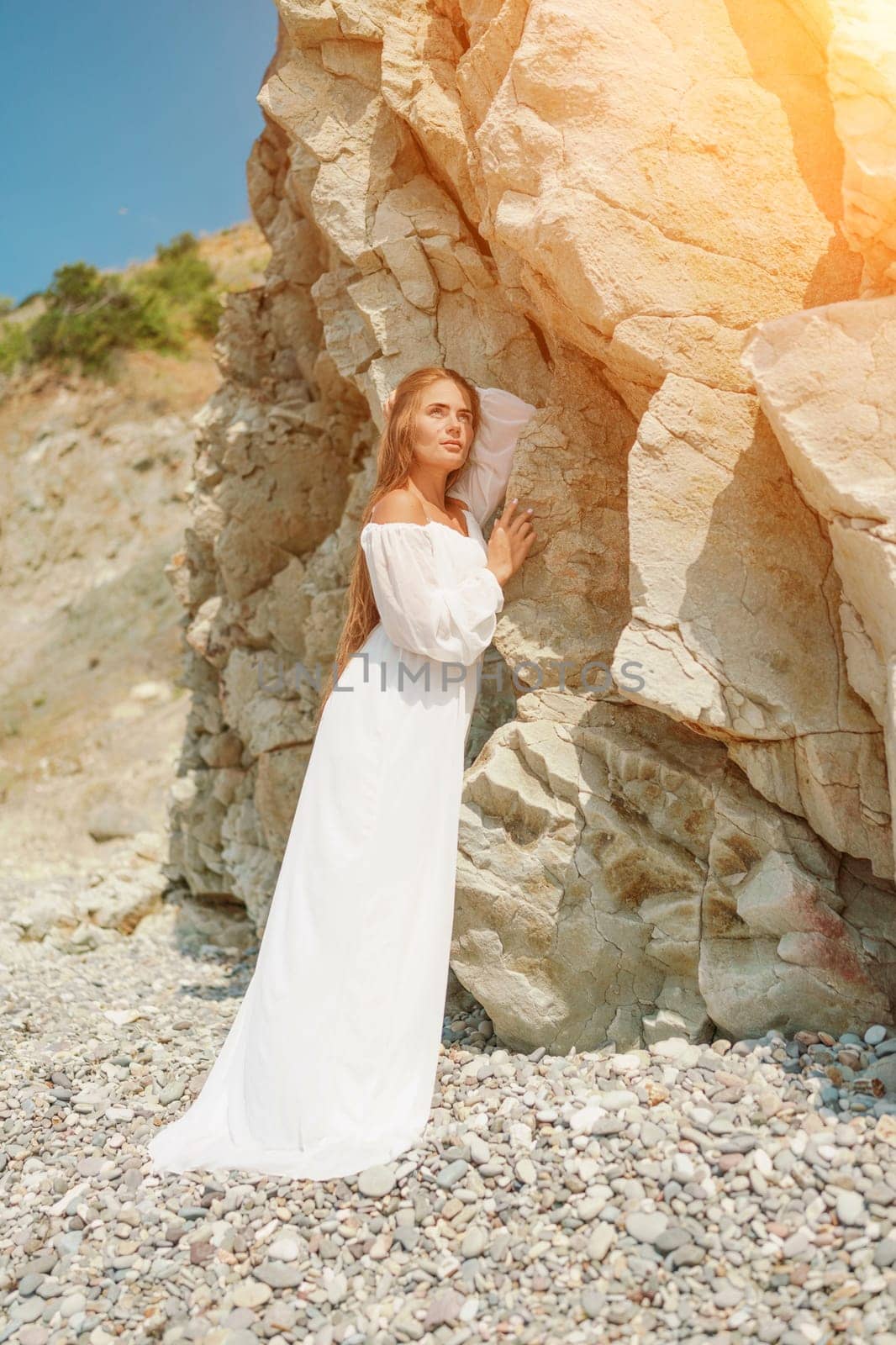 A woman in a white dress is standing on a rocky beach. Concept of serenity and tranquility, as the woman is enjoying the natural beauty of the beach