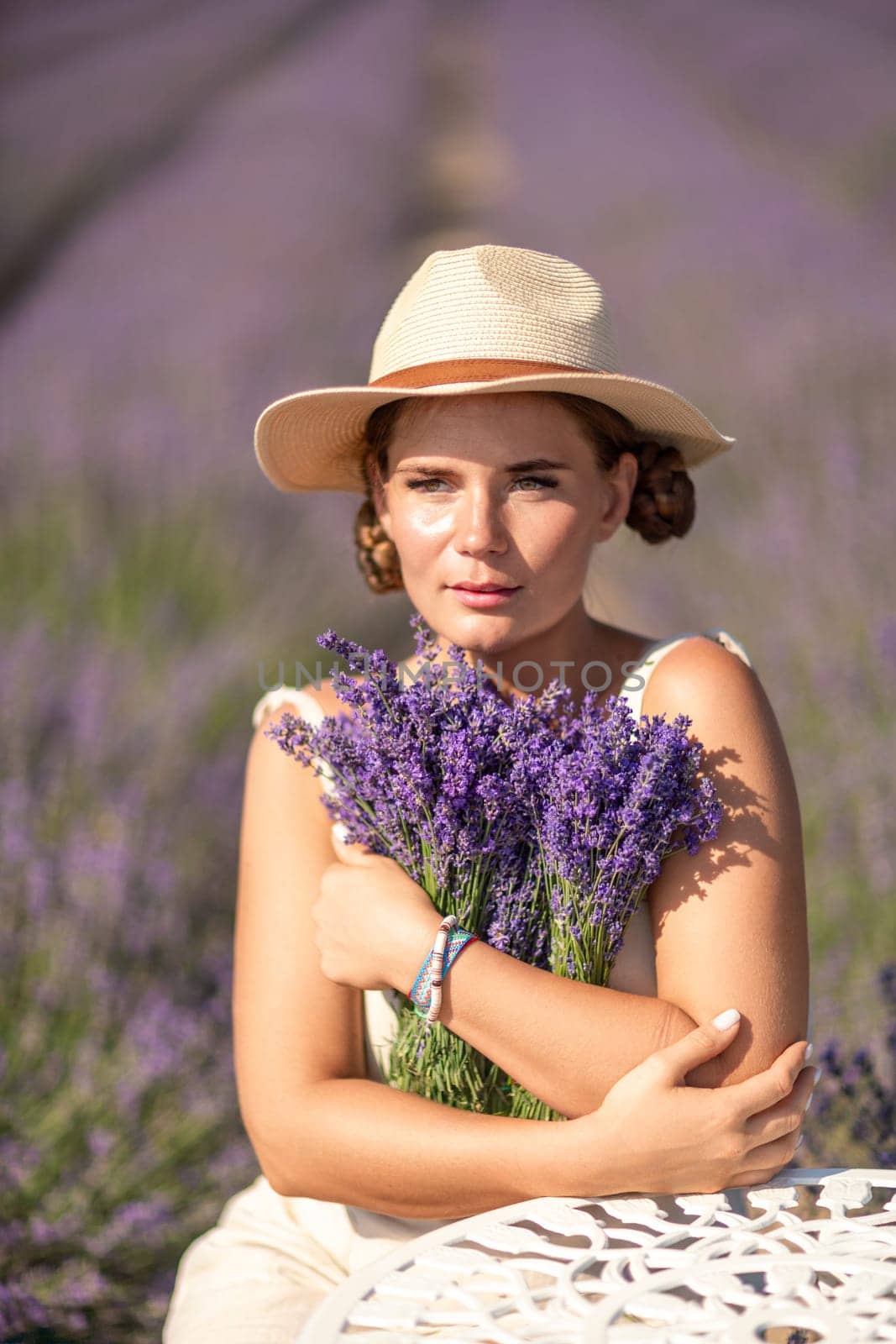 A woman is sitting in a field of lavender flowers and wearing a straw hat. She is smiling and holding a bouquet of flowers. Scene is peaceful and serene, as the woman is surrounded by the beauty of nature.