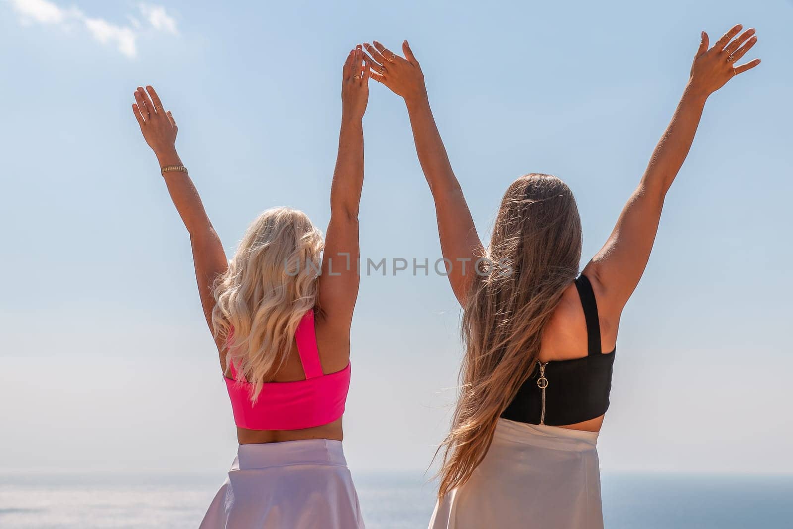 Two women are standing on a beach, one wearing a pink top and the other wearing a black top. They are both holding their hands up in the air, as if they are celebrating something. The sky is clear