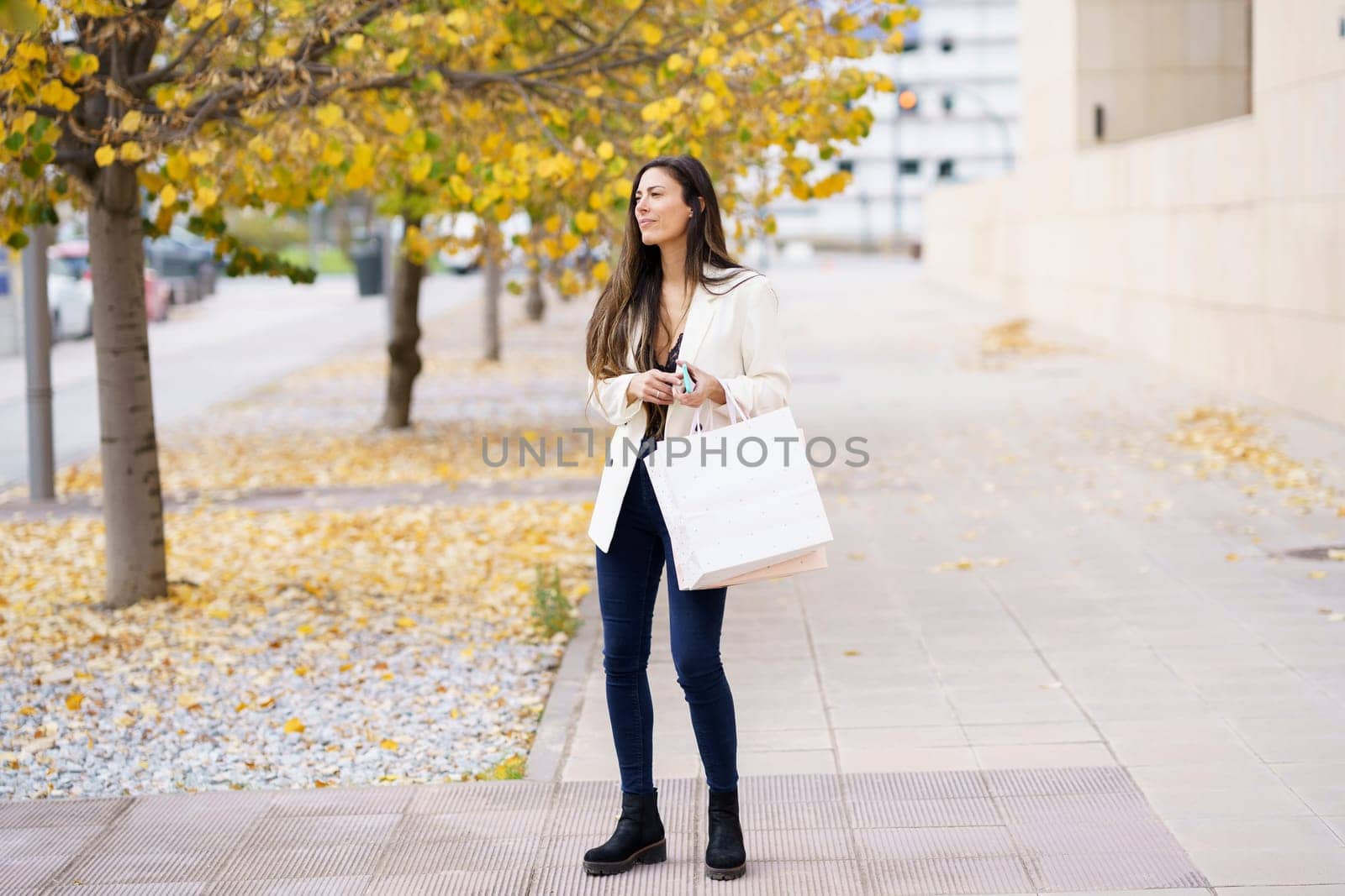 A stylish young woman walks with poise and grace down a beautiful, picturesque street adorned with colorful autumn leaves, joyfully holding a shopping bag as she enjoys her day out exploring