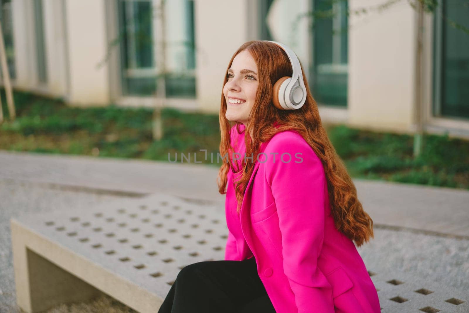 On a beautiful day, a vibrant young woman with long red hair enjoys music outdoors. Wearing stylish headphones and a bright pink jacket, she embodies carefree enjoyment and a casual lifestyle