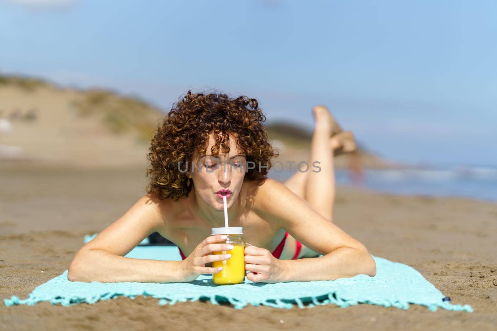 Content curly lady in bikini sunbathing on coastline and drinking fresh orange juice from cup in sunlight