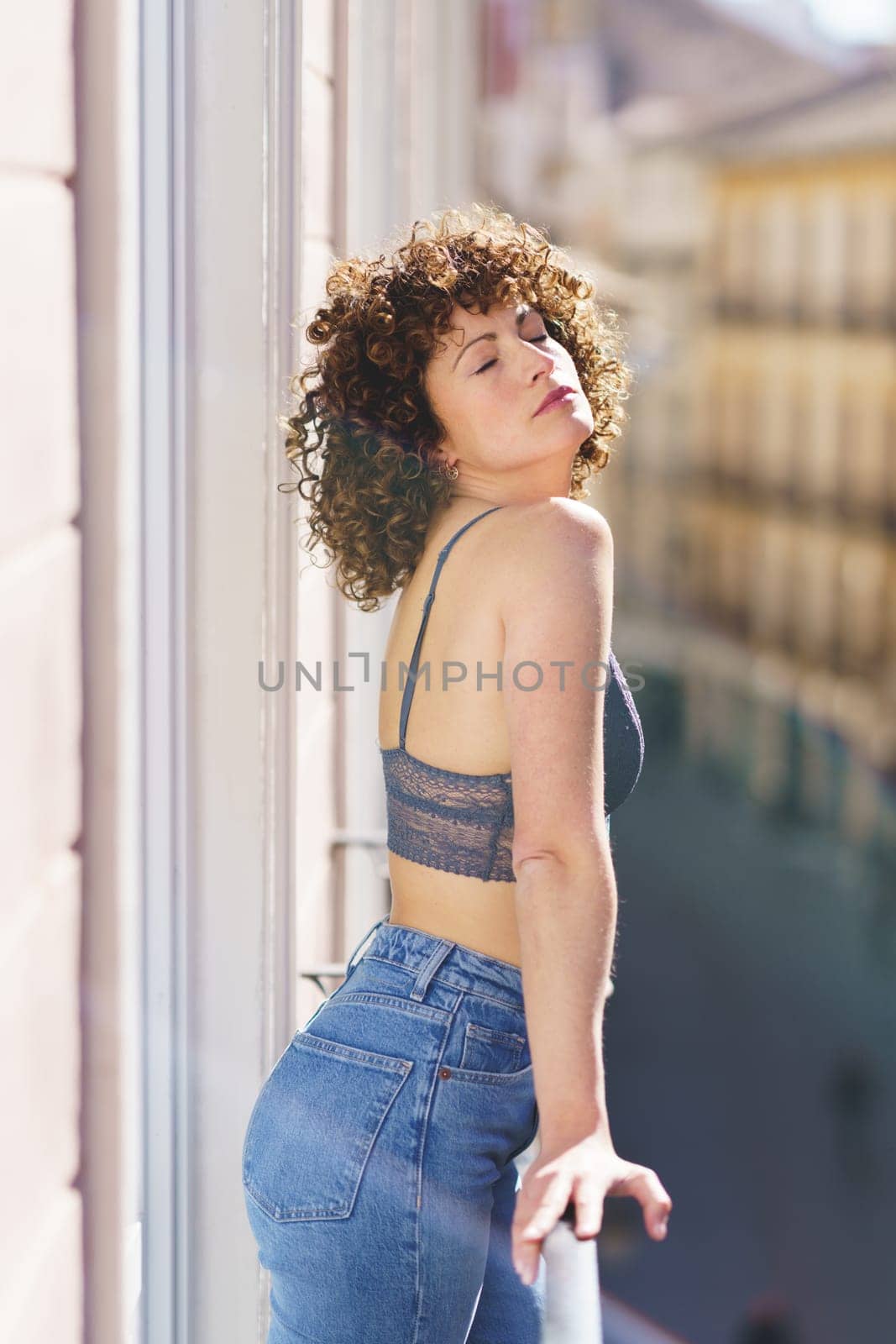 Calm young female in bra and jeans with Afro hairstyle standing on balcony with closed eyes while leaning on fence on sunny day
