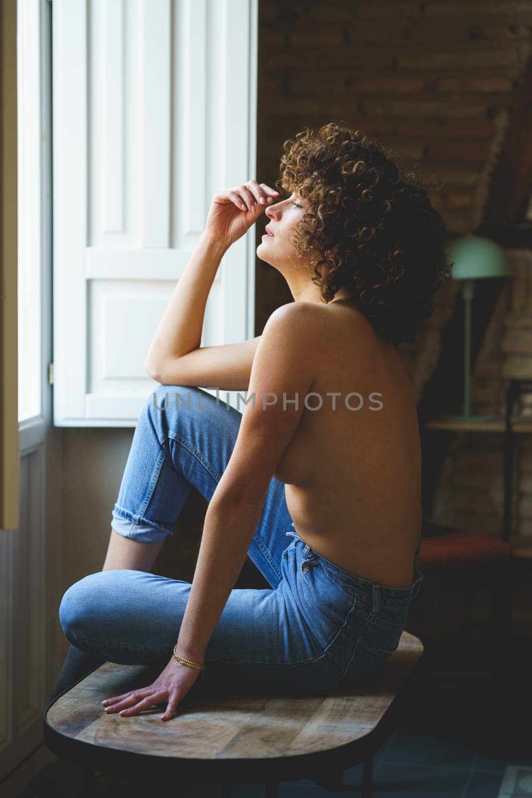 Side view of sensual young topless woman, with curly hair sitting on wooden stool with raised leg near window with shutters in room with brick wall