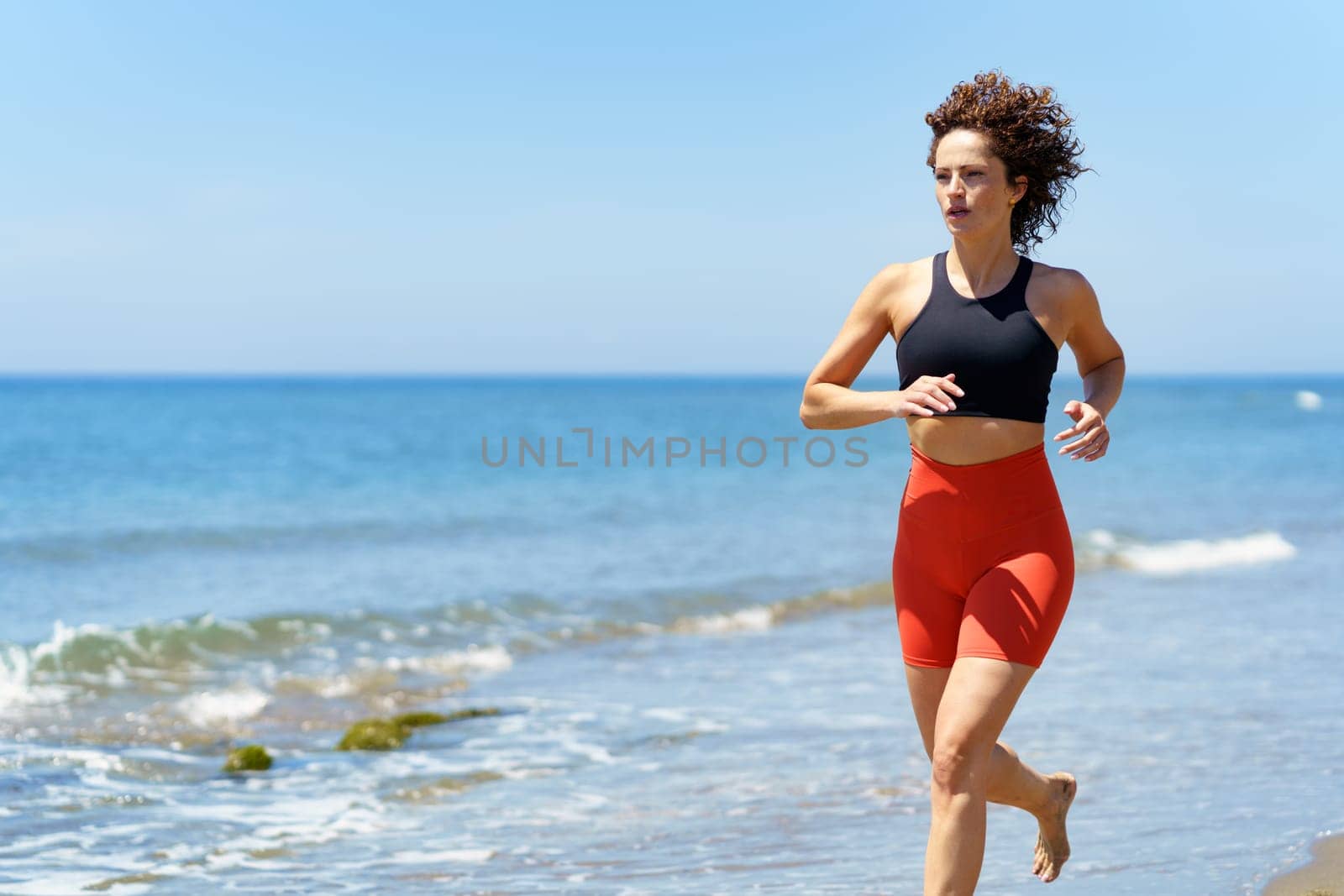 Concentrated barefoot young female with curly hair, wearing sportswear and looking away while jogging in daylight on sandy beach near wavy seawater against blurred blue sky