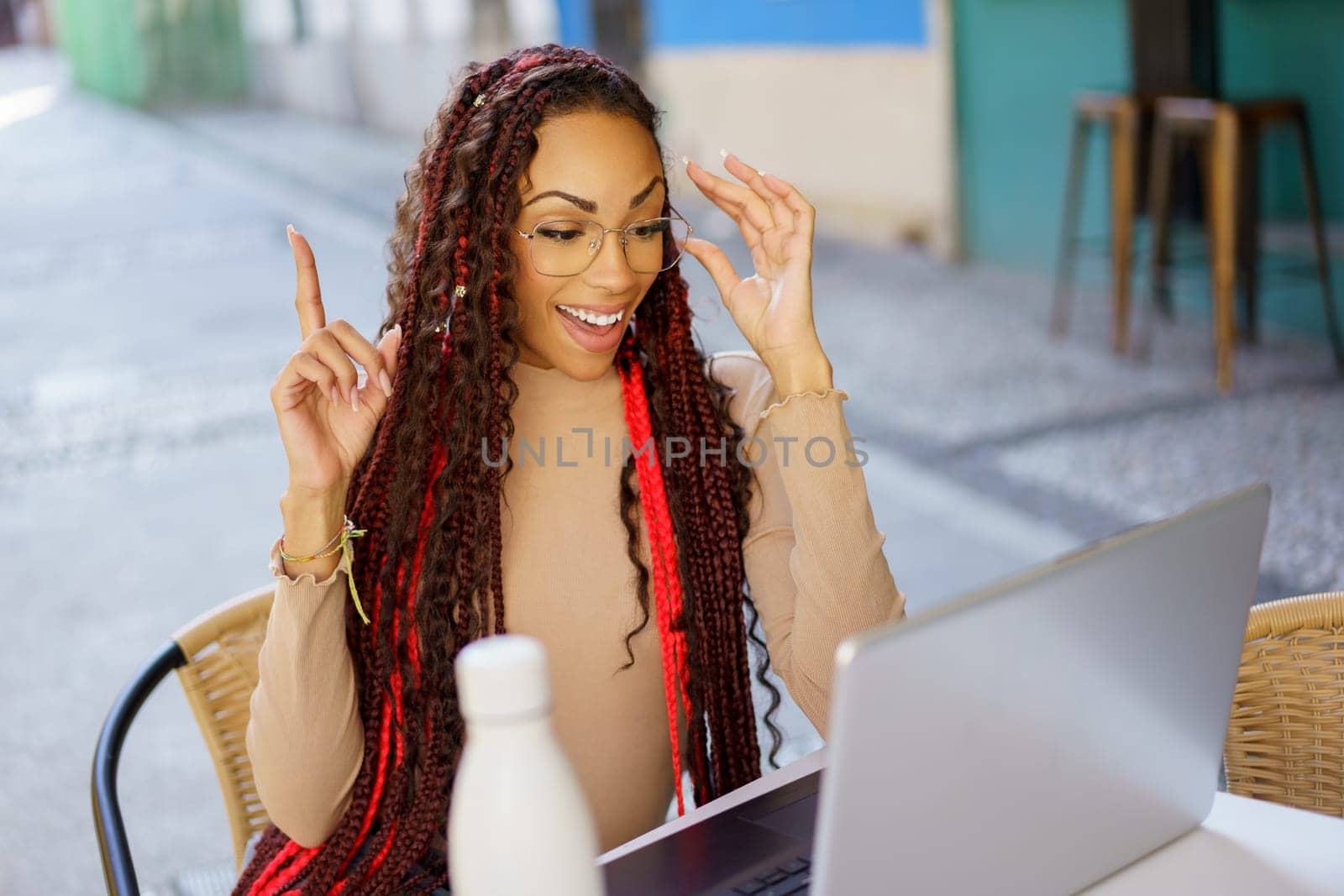 A joyful and enthusiastic young woman with beautifully braided hair, expresses her excitement while using a sleek laptop in a cozy and charming cafe setting that radiates warmth and comfort