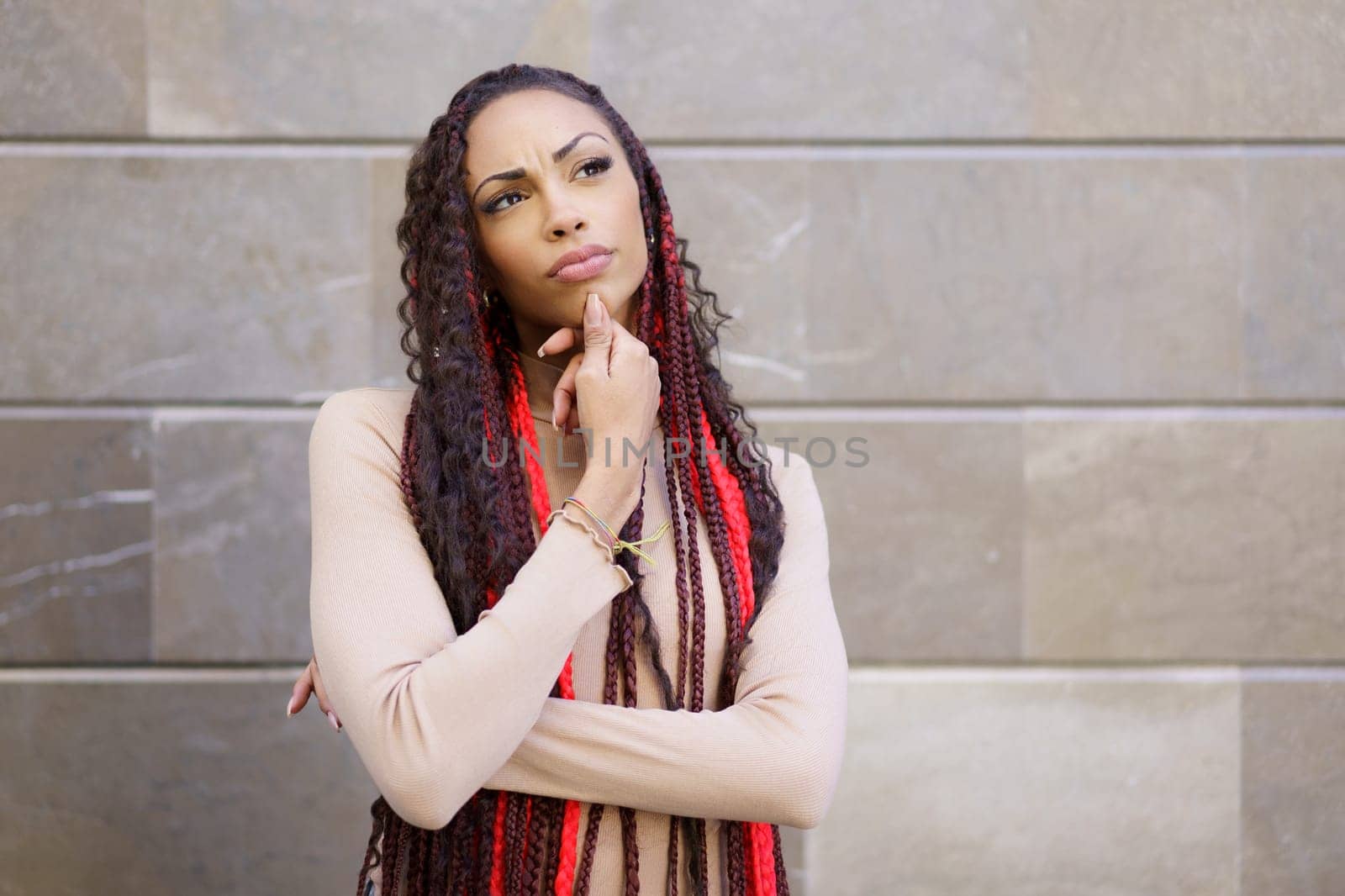 A woman stands thoughtfully against an intriguing textured wall, showcasing her distinctive braided hairstyle adorned with vibrant red accents, embodying confidence, creativity, and personal style