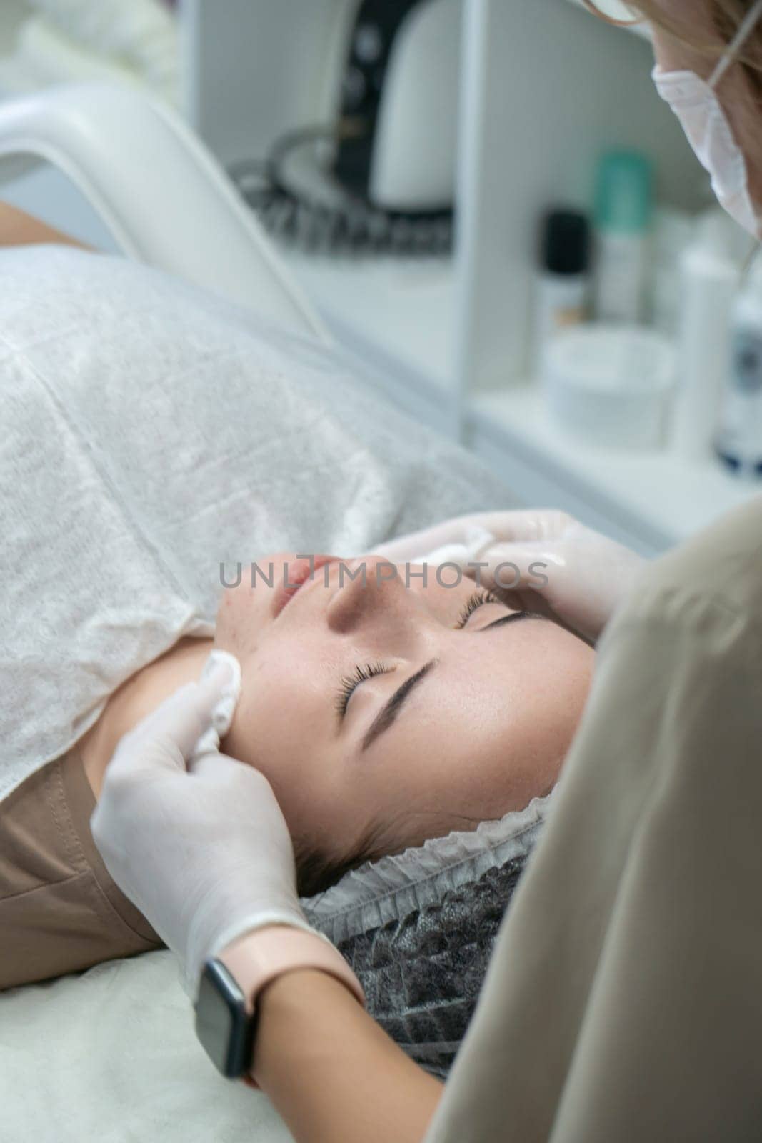Close-up of a woman receiving a facial treatment. A beautician's gloved hands gently cleanse her face. Relaxed client lies on a spa bed.