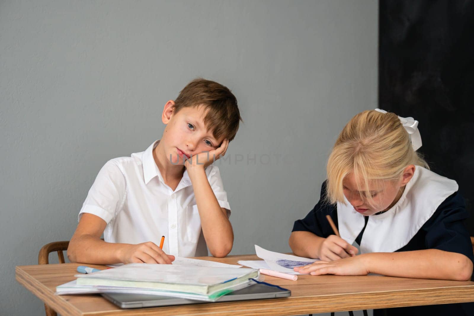 A bored boy rests his head on his hand while a girl diligently writes in a notebook at a wooden desk.