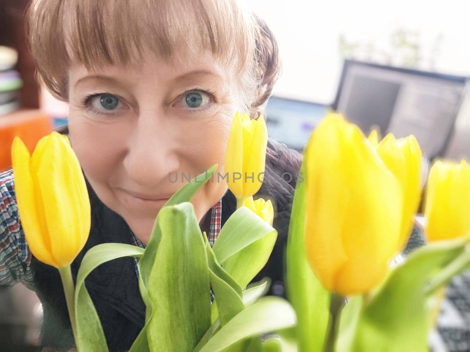 A woman with bright blue eyes smiles warmly while holding a bouquet of fresh yellow tulips in a lively indoor environment.