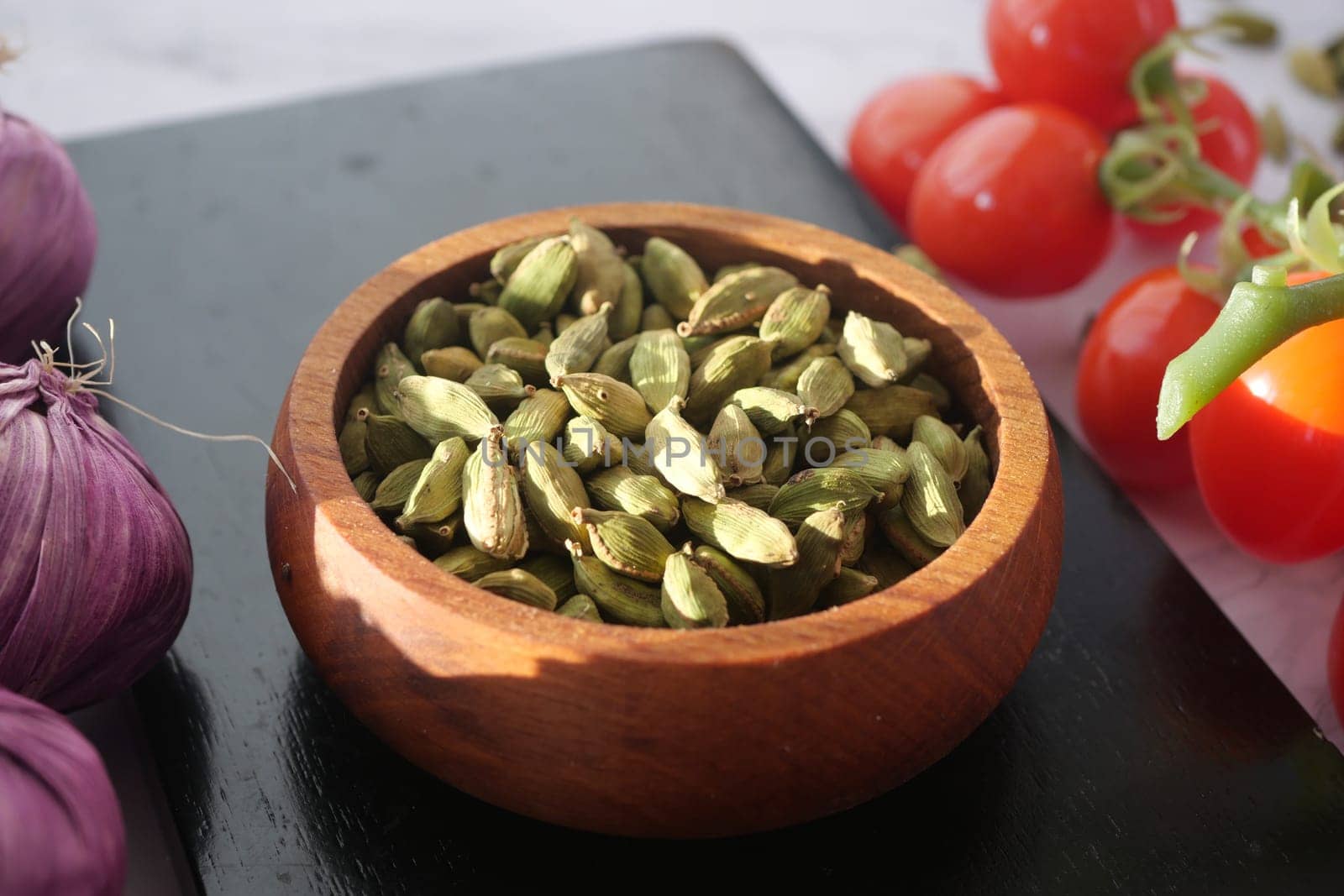 Fresh cardamom pods rest in a wooden bowl alongside cherry tomatoes.