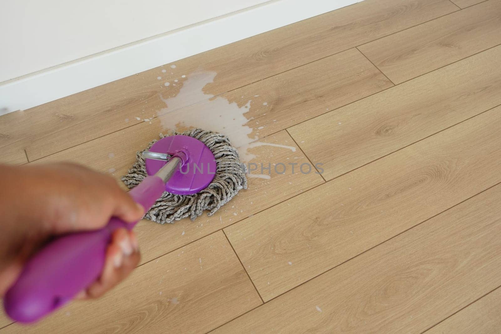 A person mops a light wooden floor with a purple mop in a sunny space.